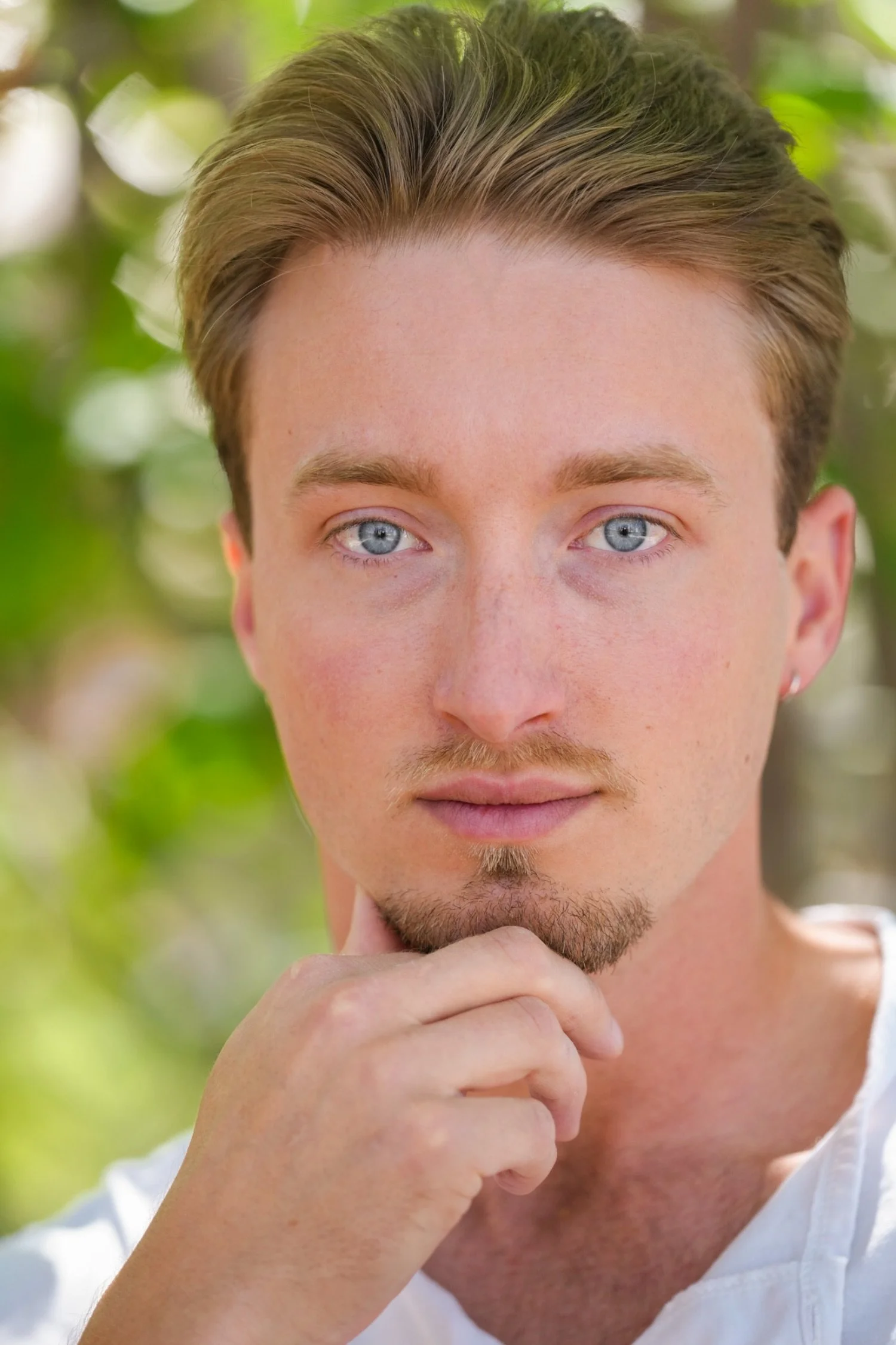 Close-up of a young man with blue eyes, light brown hair, and a trimmed beard, thoughtfully resting his chin on his hand against a blurred green foliage background.