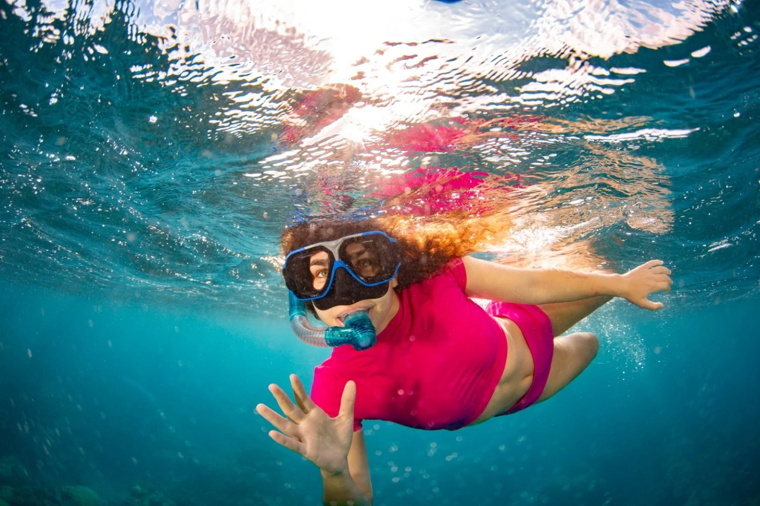A woman snorkeling underwater, wearing a black mask, blue snorkel, and a pink top and shorts, waving at the camera.