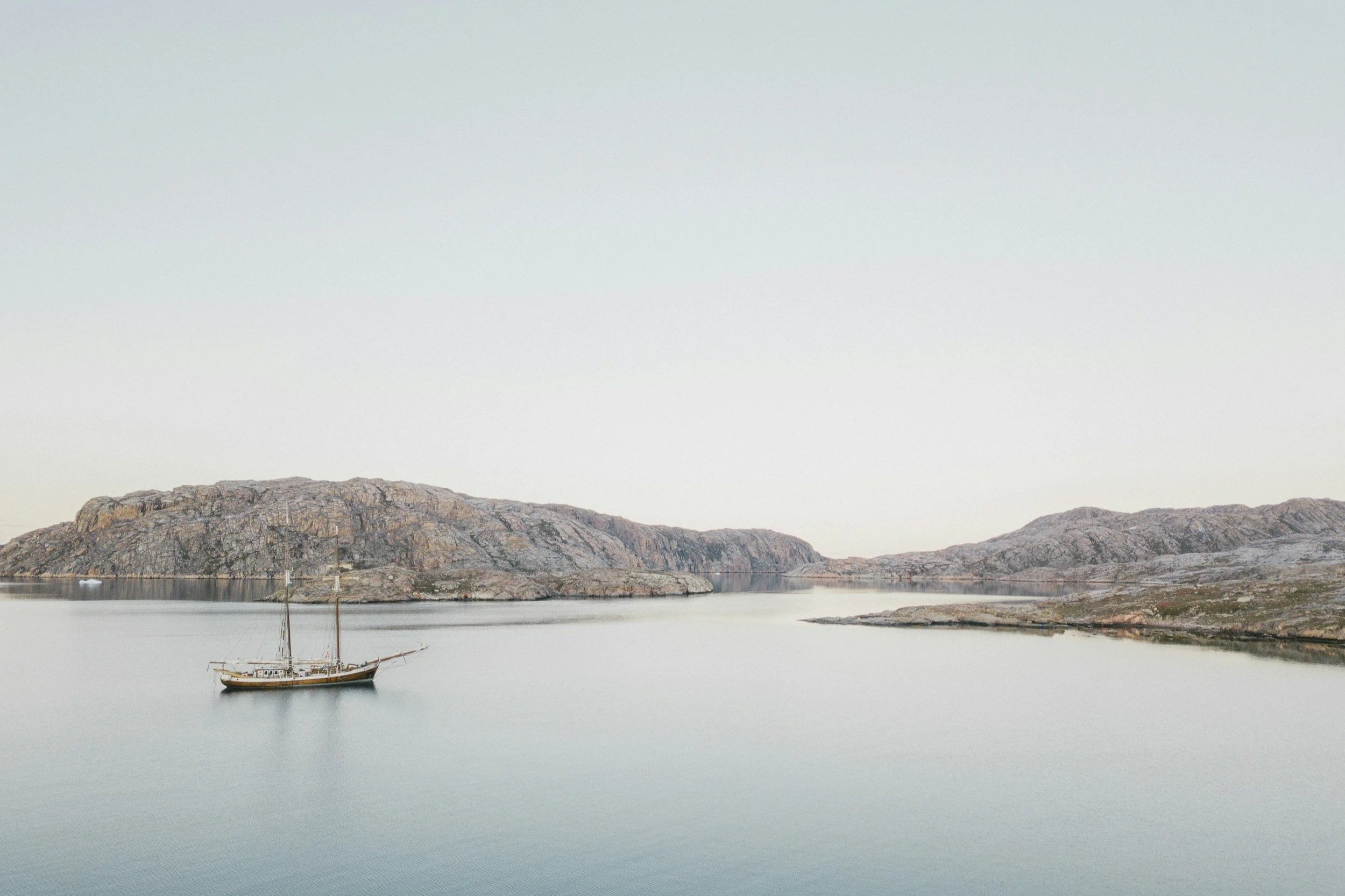 A sailboat floats on calm water near rocky islands or hills under a pale sky.