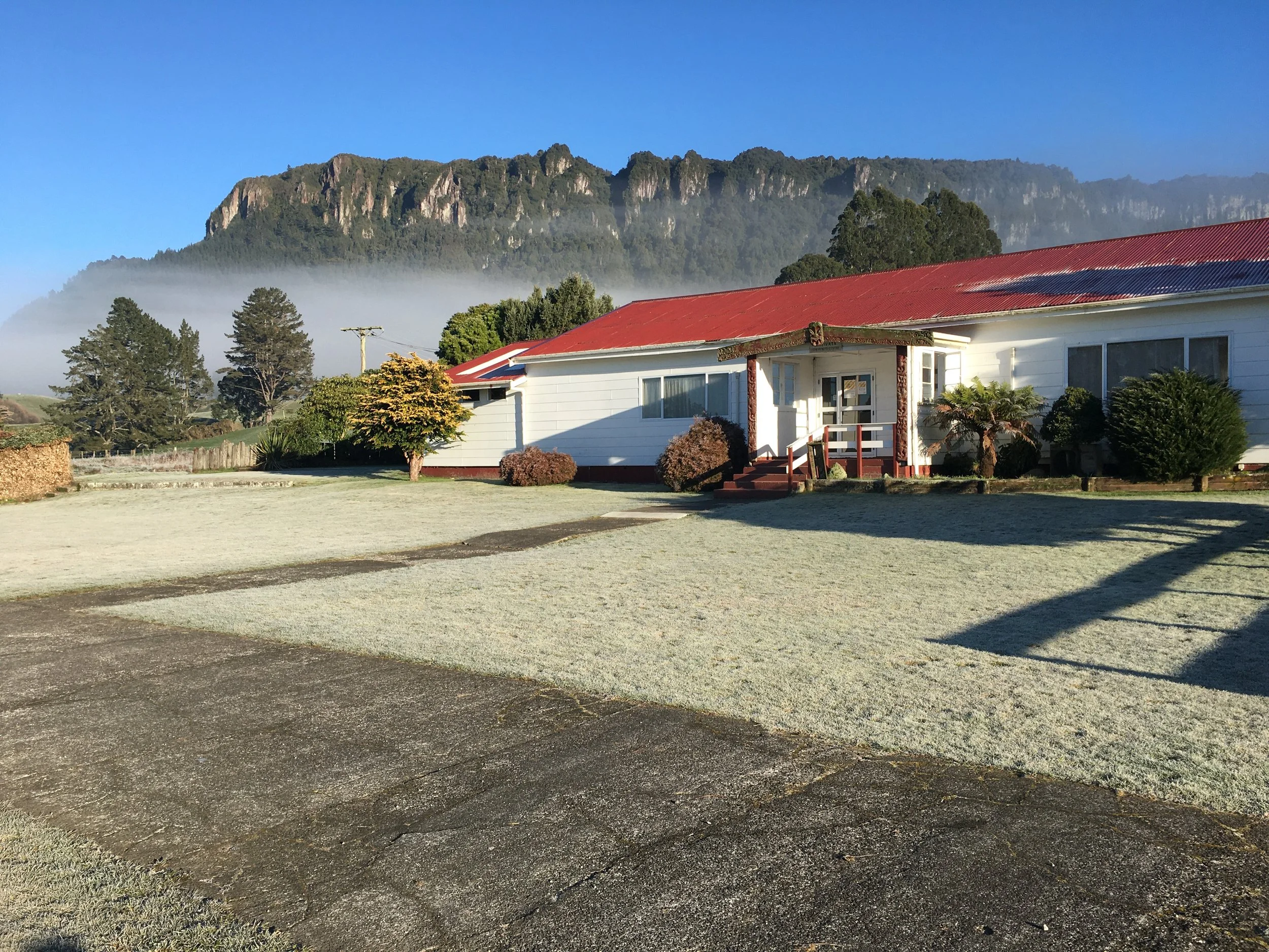 A white wharekai with a red roof, surrounded by grass and trees, with mountains and mist in the background.