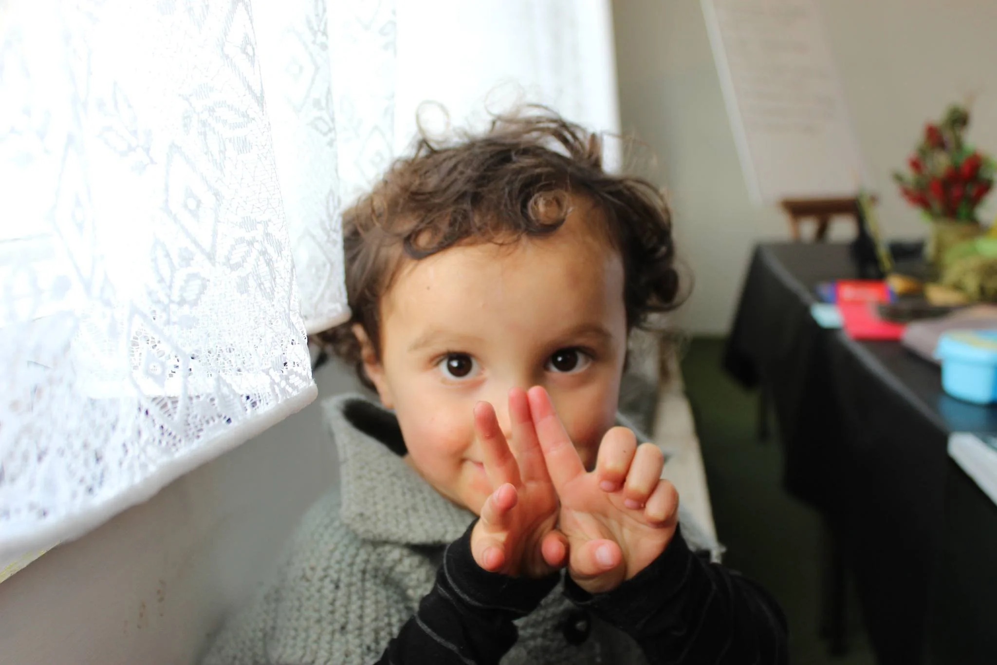 A young child with curly brown hair and big eyes looking at the camera while holding hands near their face.