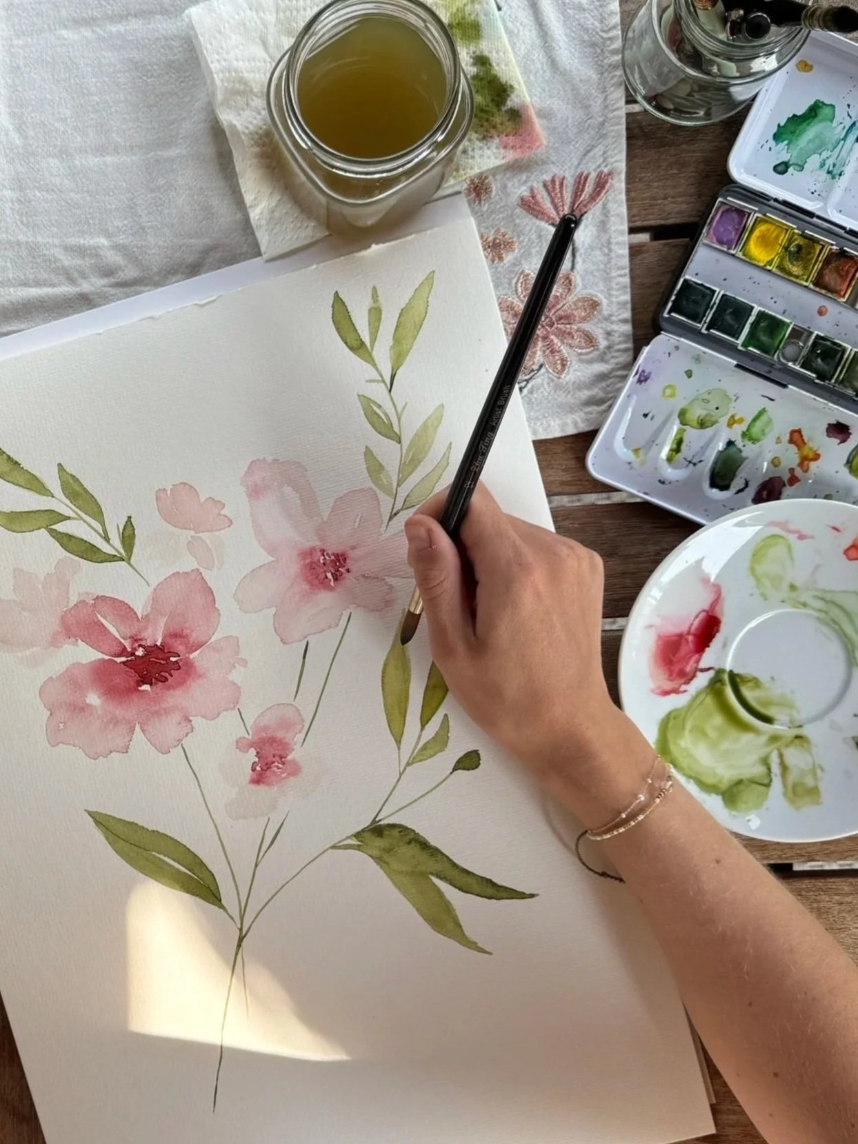 Eine Person malt mit Wasserfarben ein Blumenbild auf Papier, umgeben von Malutensilien und einer Tasse mit grünlicher Flüssigkeit.