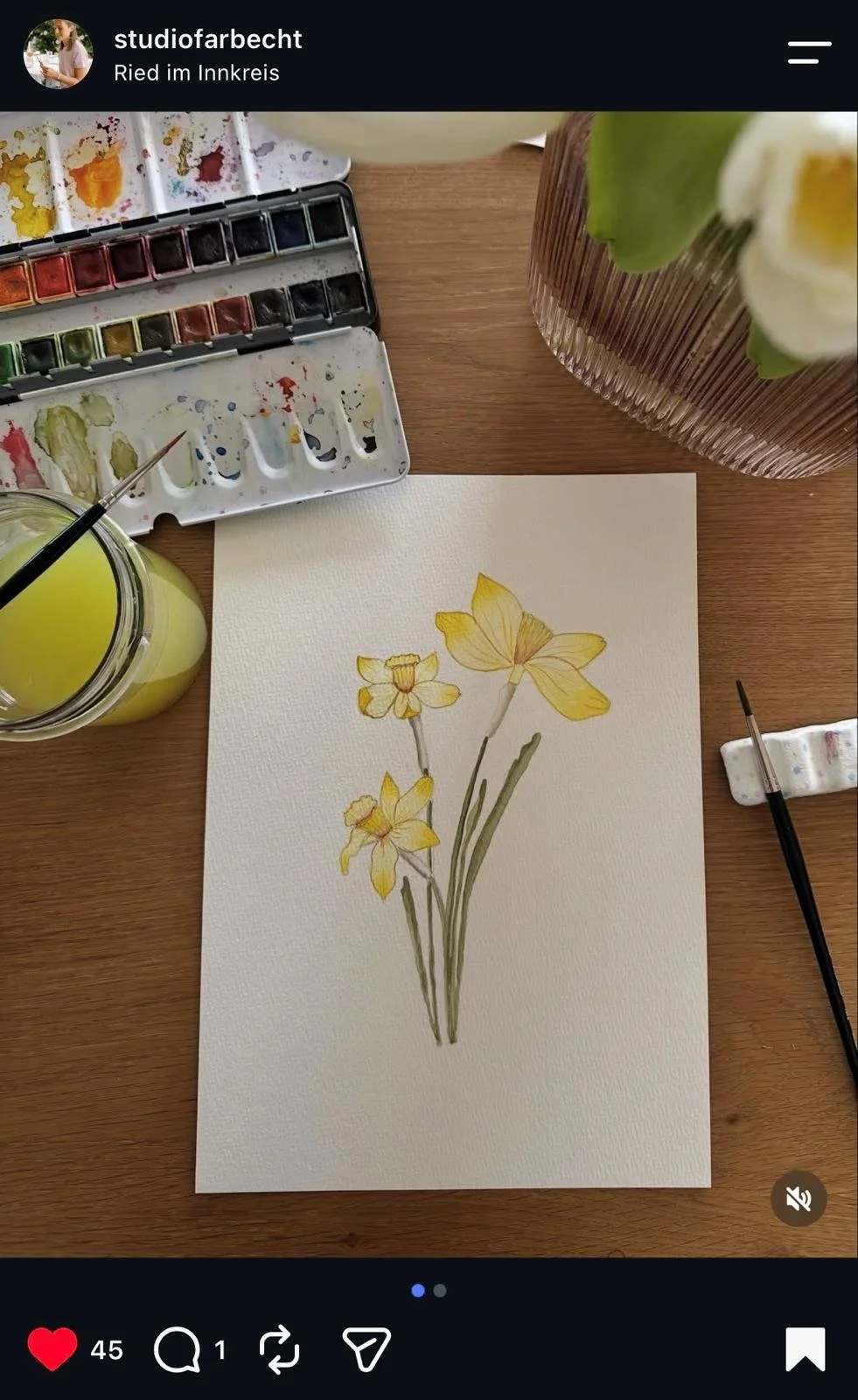 A watercolor painting of yellow daffodils on a white textured paper, surrounded by watercolor paint set, brushes, and a jar of yellow-green liquid, on a wooden table.