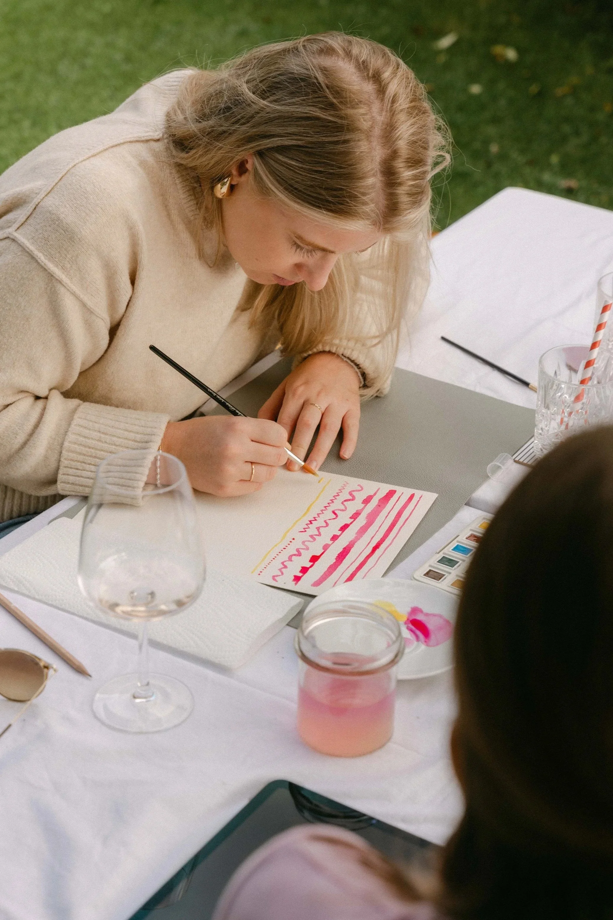 Eine Frau bemalt ein Papier mit pinken, roten und gelben Mustern auf einem Tisch im Freien.