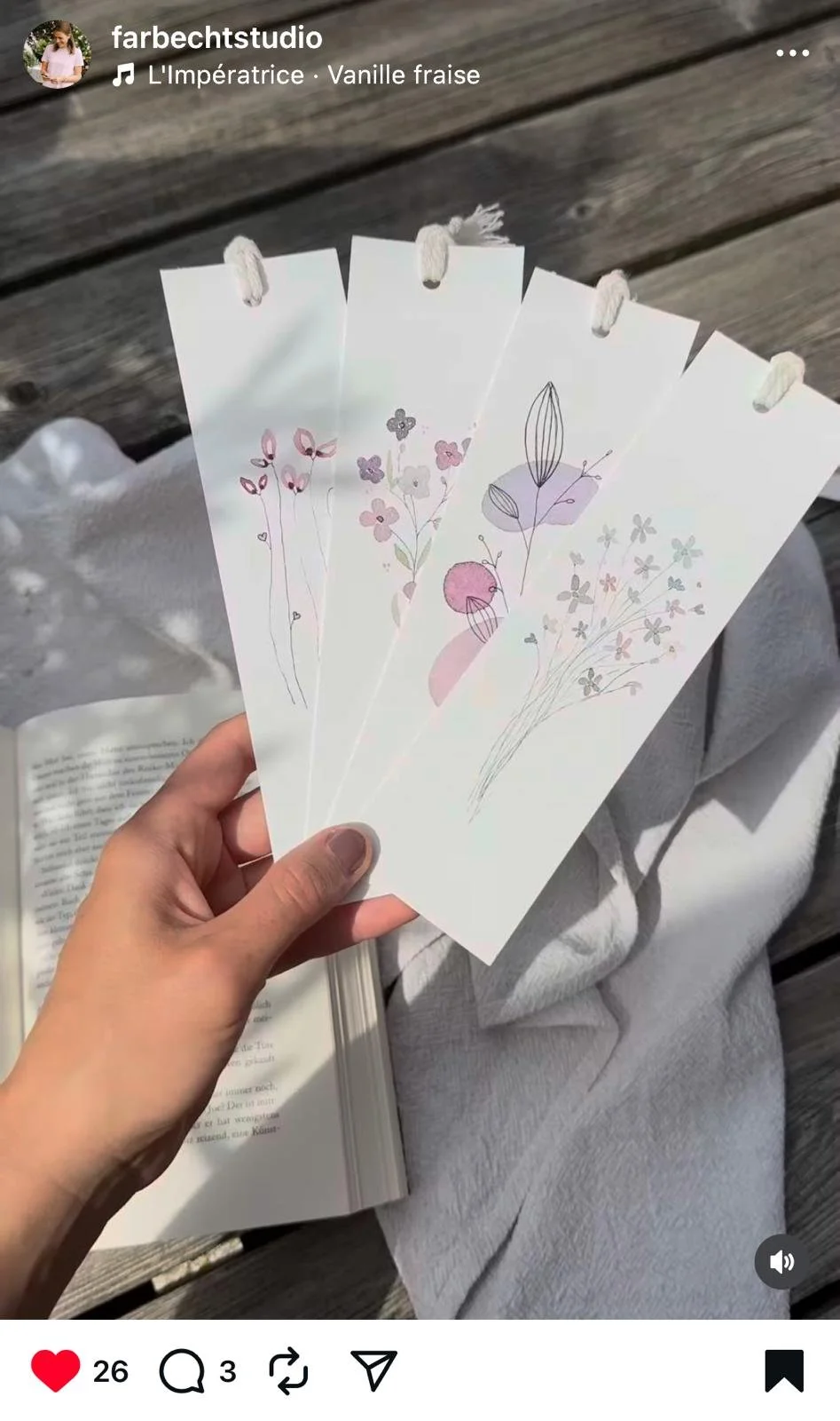 Hand hält drei zarte, minimalistische Postkarten mit blumigen Illustrationen in Pastellfarben. Im Hintergrund liegt ein aufgeschlagenes Buch, und die Person sitzt auf Holz.