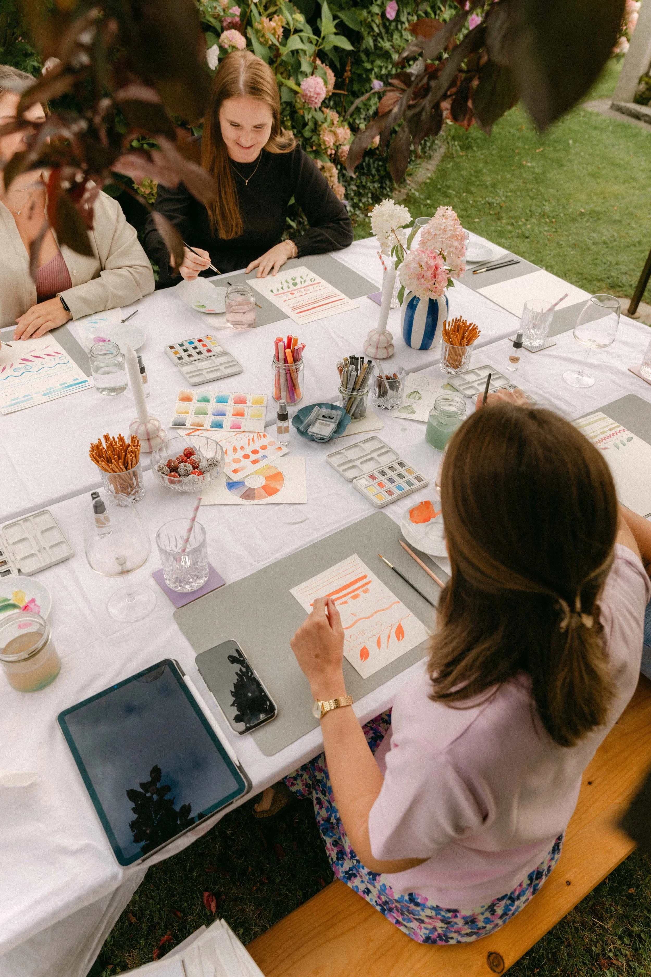 Mehrere Frauen beim Basteln im Freien, umgeben von Blumen, mit Wasserfarben, Pinsel und Bastelmaterial auf einem weißen Tisch.