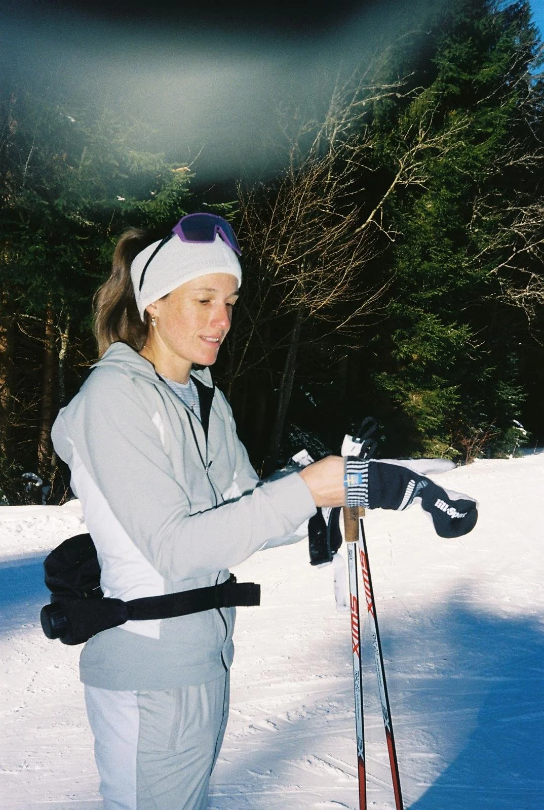Frau in Winterkleidung mit Skiern und Skistöcken im Schnee, im Hintergrund Bäume, sonniger Tag.