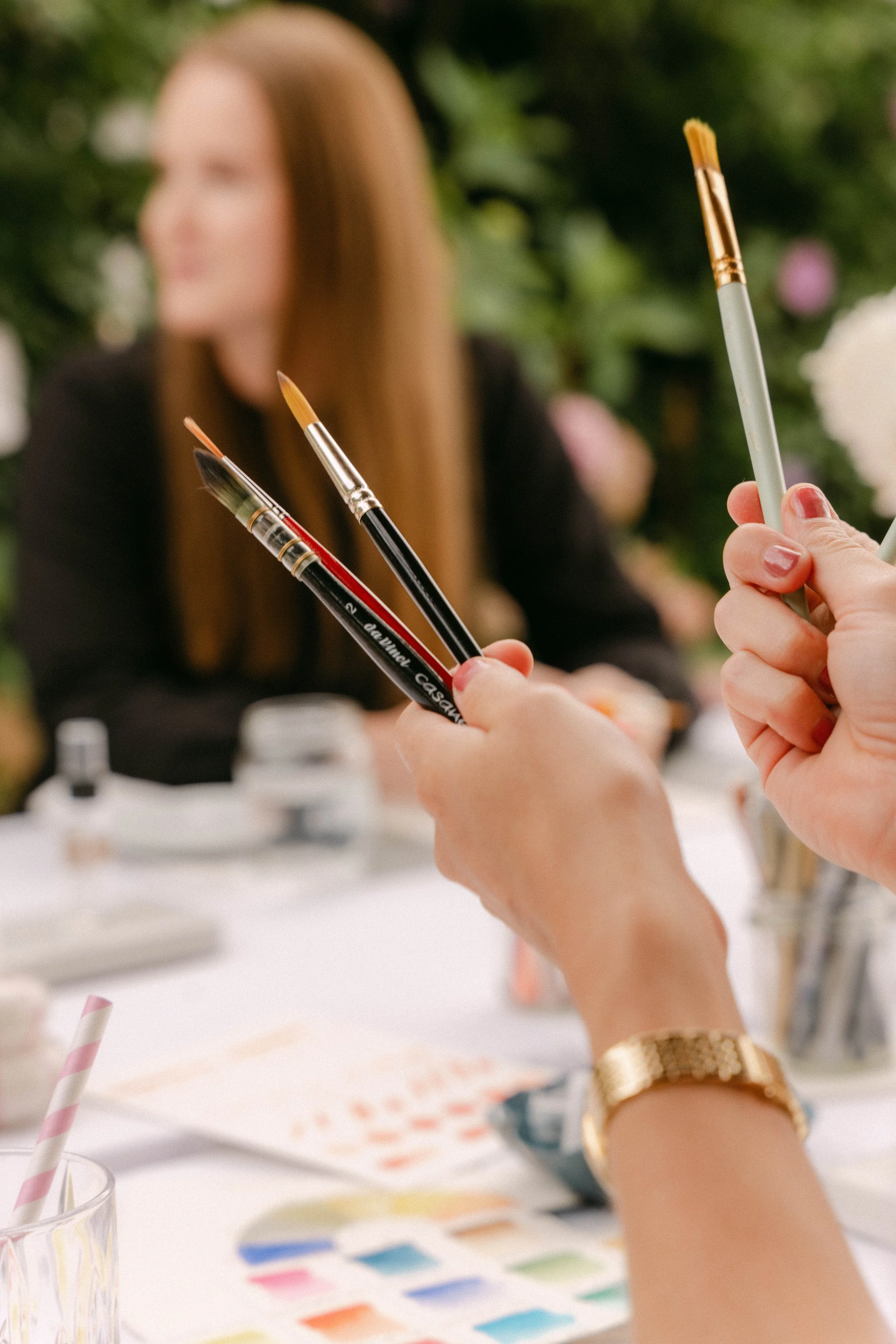Hand hält Pinsel und Farbstifte bei einem Mal-Workshop in einem Blumengarten, mit einer Frau im Hintergrund.