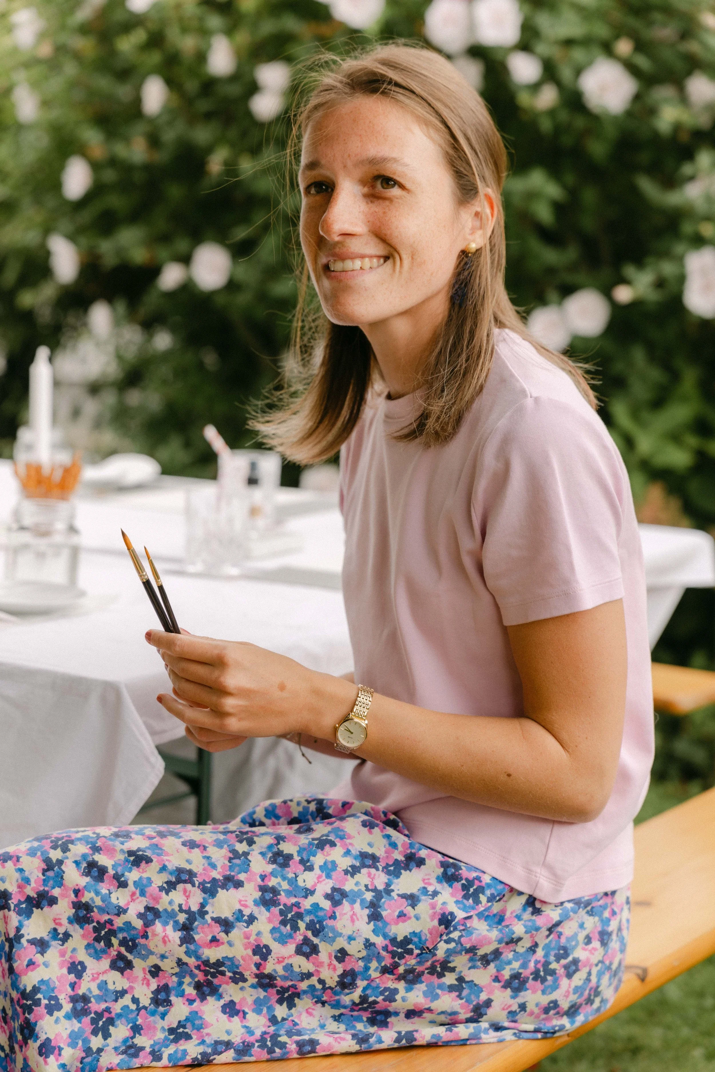 Junge Frau mit geblümtem Rock und rosa T-Shirt sitzt auf einer Bank im Garten, hält Pinsel in den Händen, vor ihr ein weißer Tisch mit Malutensilien, im Hintergrund blühende Pflanzen.