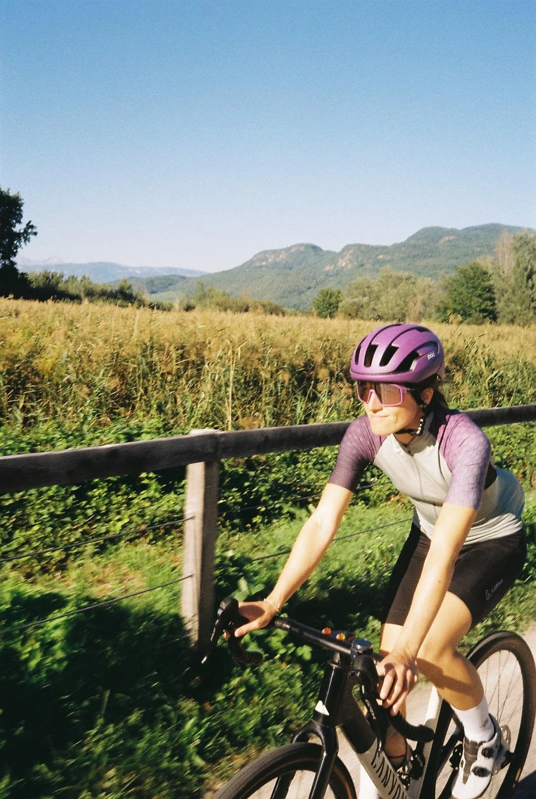 Frau fährt Fahrrad auf Landstraße, umgeben von Feldern, Bäumen und Bergen im Hintergrund, sonniger Tag, trägt lilafarbenen Helm und Sonnenbrille.