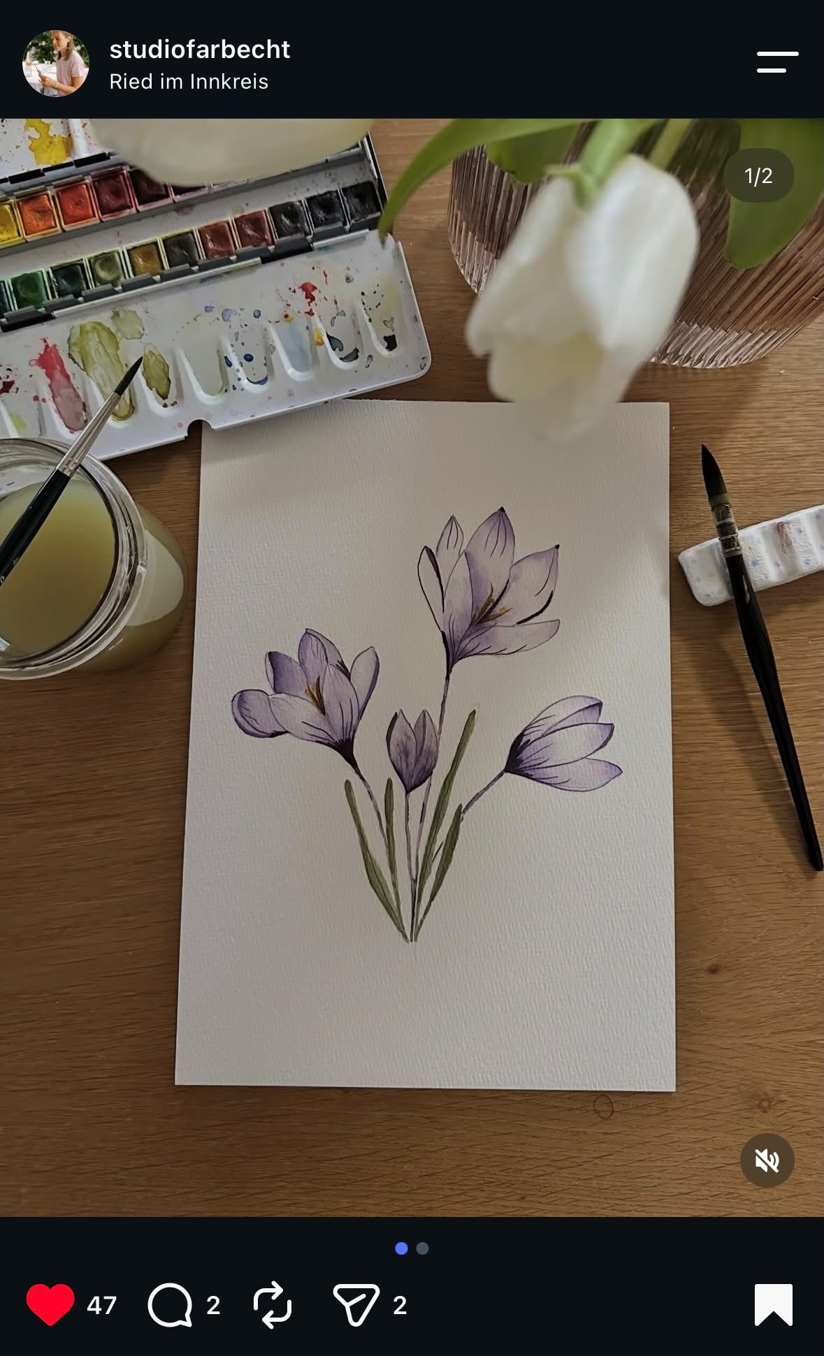 A watercolor painting of purple crocus flowers on a white sheet of paper, surrounded by painting supplies, including watercolors, paintbrushes, and a jar of water, on a wooden table.