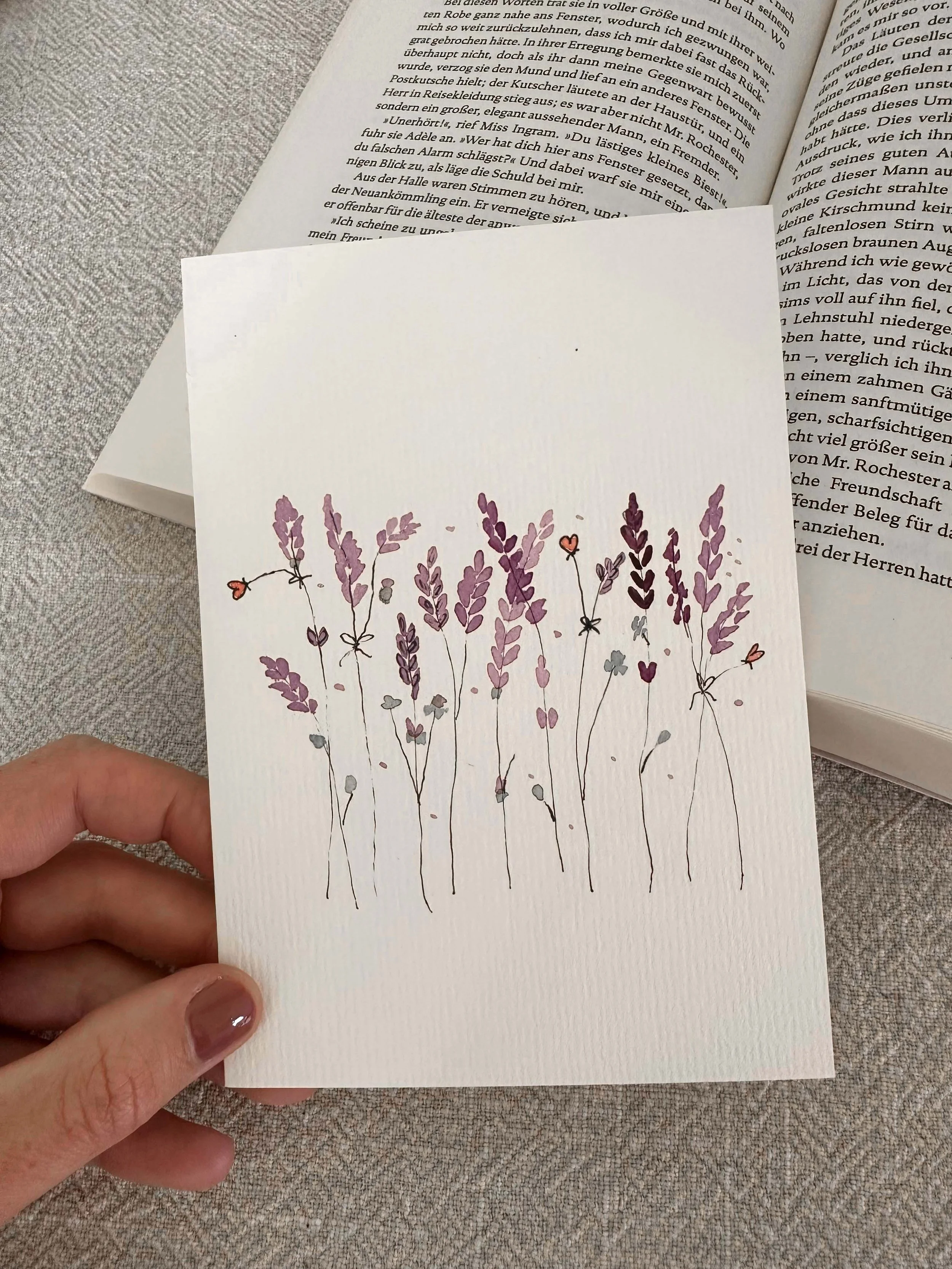Eine handgehaltene Karte mit einer Illustration von lilafarbenen Blumen und kleinen roten Herzmotiven auf weißem Hintergrund. Im Hintergrund sind aufgeschlagene Bücher mit deutschem Text sichtbar.