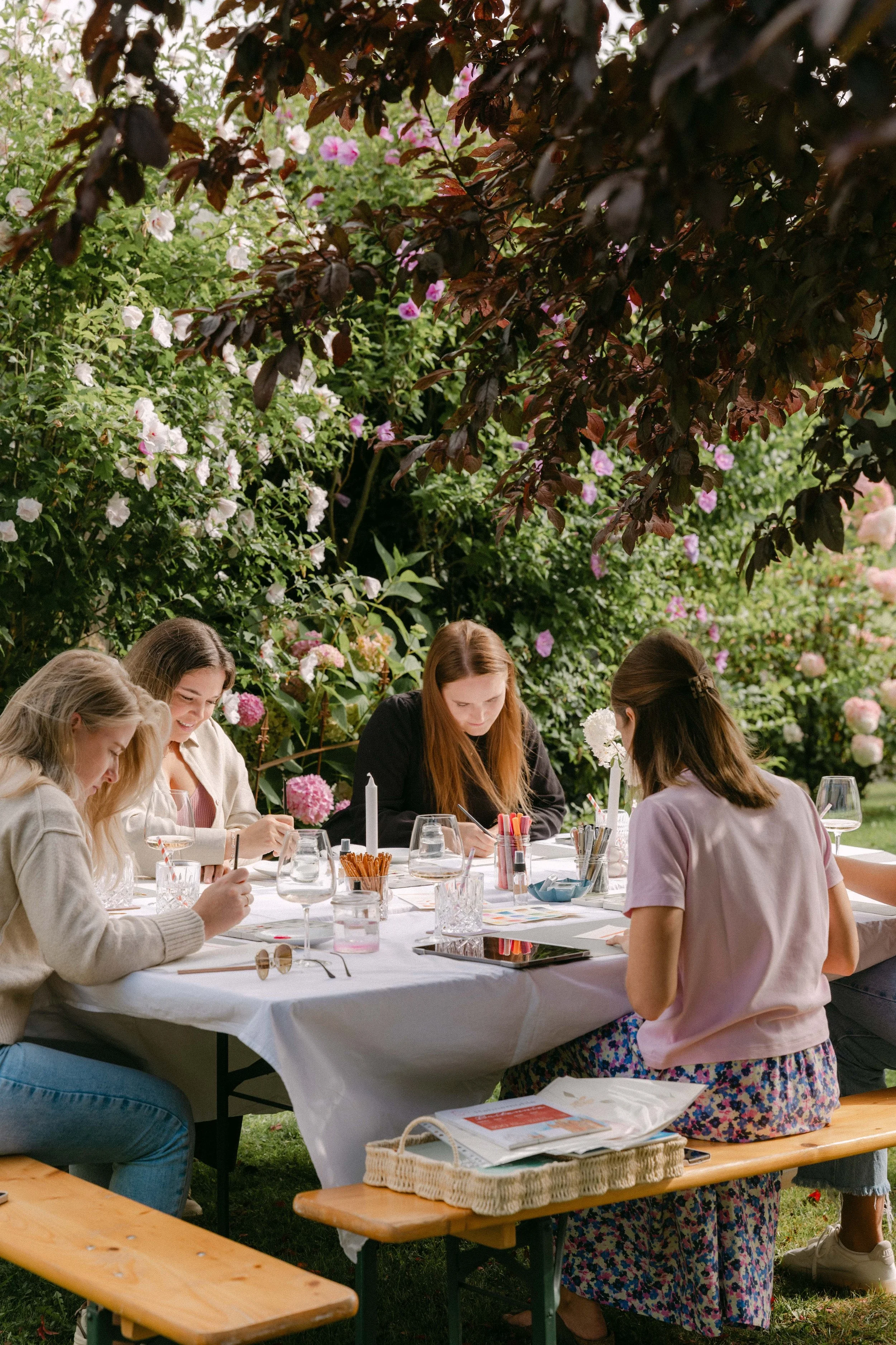 Fünf Frauen sitzen um einen Tisch im Garten, bei einer Kunstaktivität, umgeben von blühenden Büschen und Bäumen.