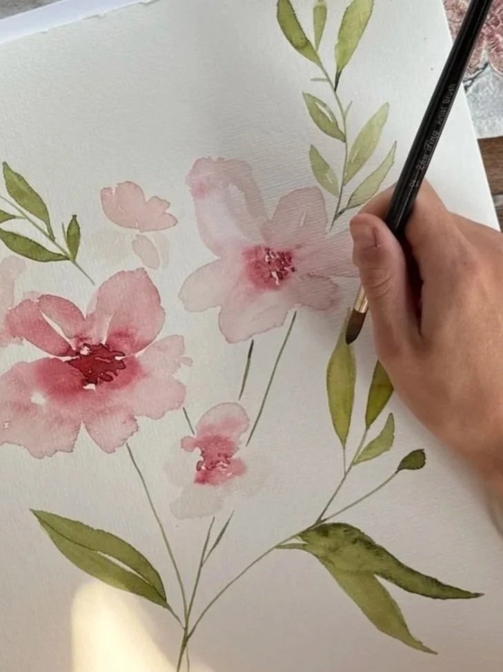 Eine Person malt mit Wasserfarben auf Papier, es sind pinkfarbene Blumen mit grünen Blättern zu sehen.