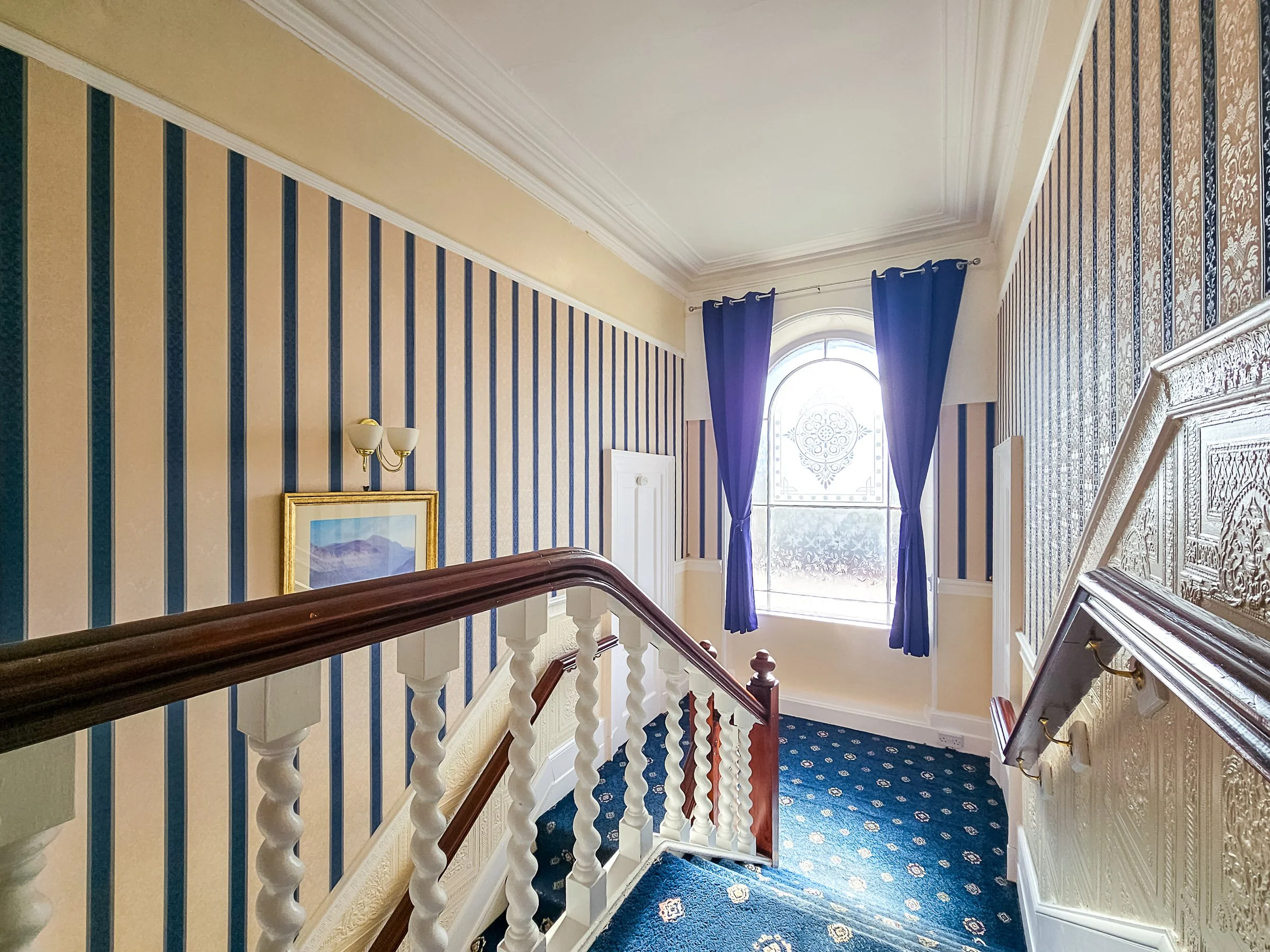 Interior view of a staircase landing with patterned wallpaper, a window with blue curtains, a framed painting, and decorative molding.
