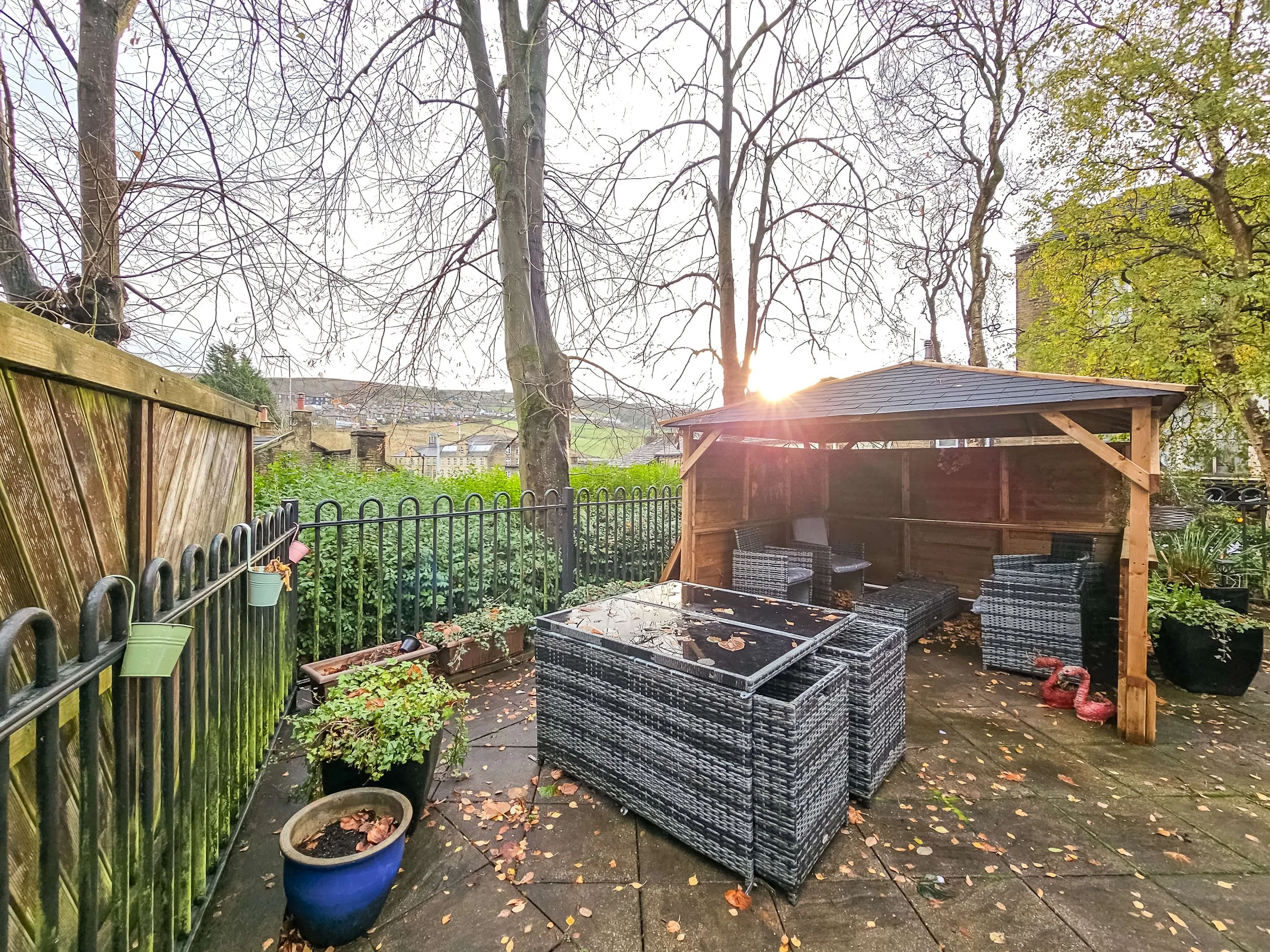 A backyard patio with outdoor furniture including a black wicker table, chairs, and a wooden shed. There are trees with bare branches, potted plants, and fallen leaves on the floor.