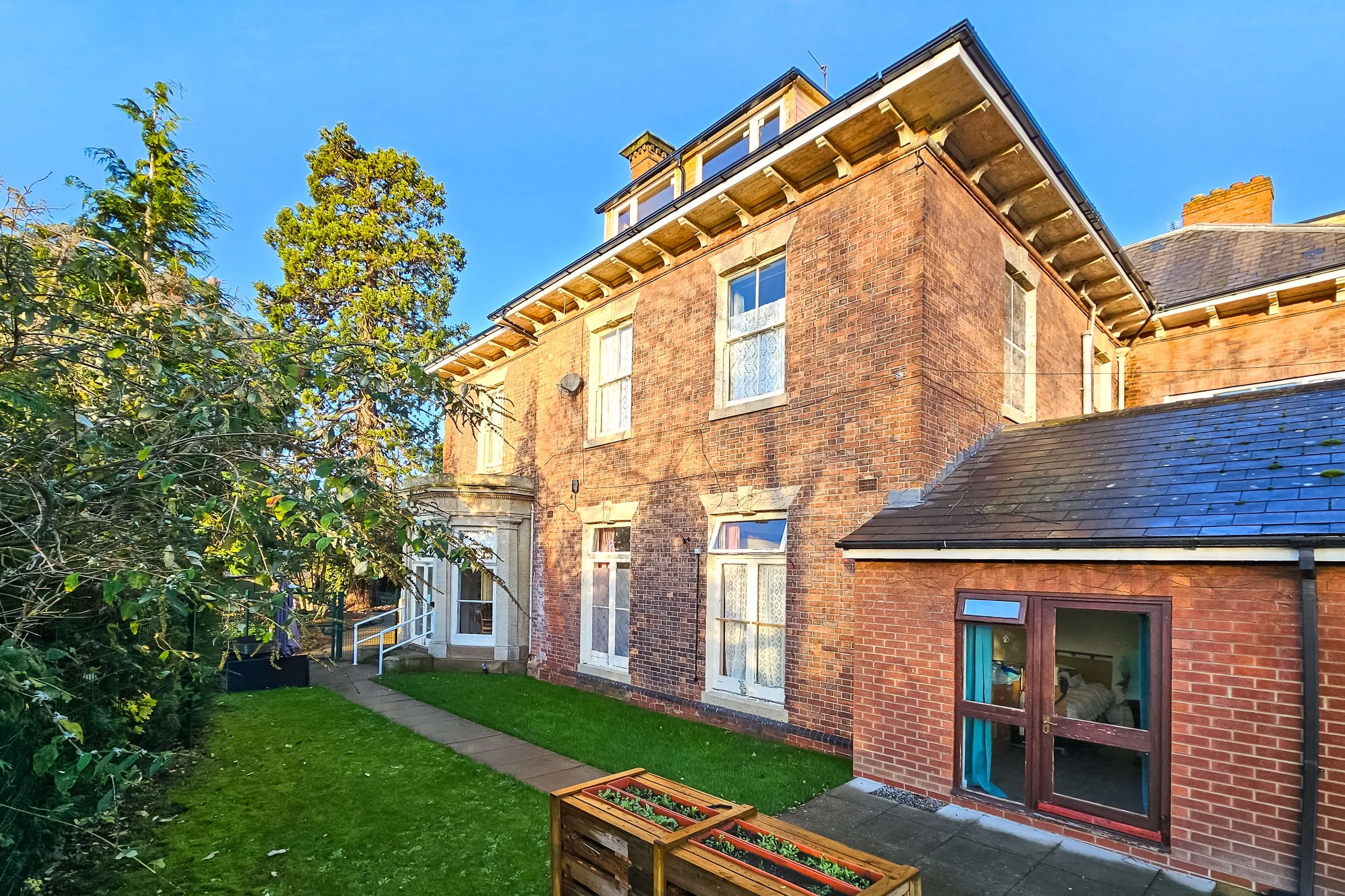 Rear view of a multi-story brick house with a small brick extension, windows with lace curtains, and a garden with a lawn, a pathway, and a flower planter.