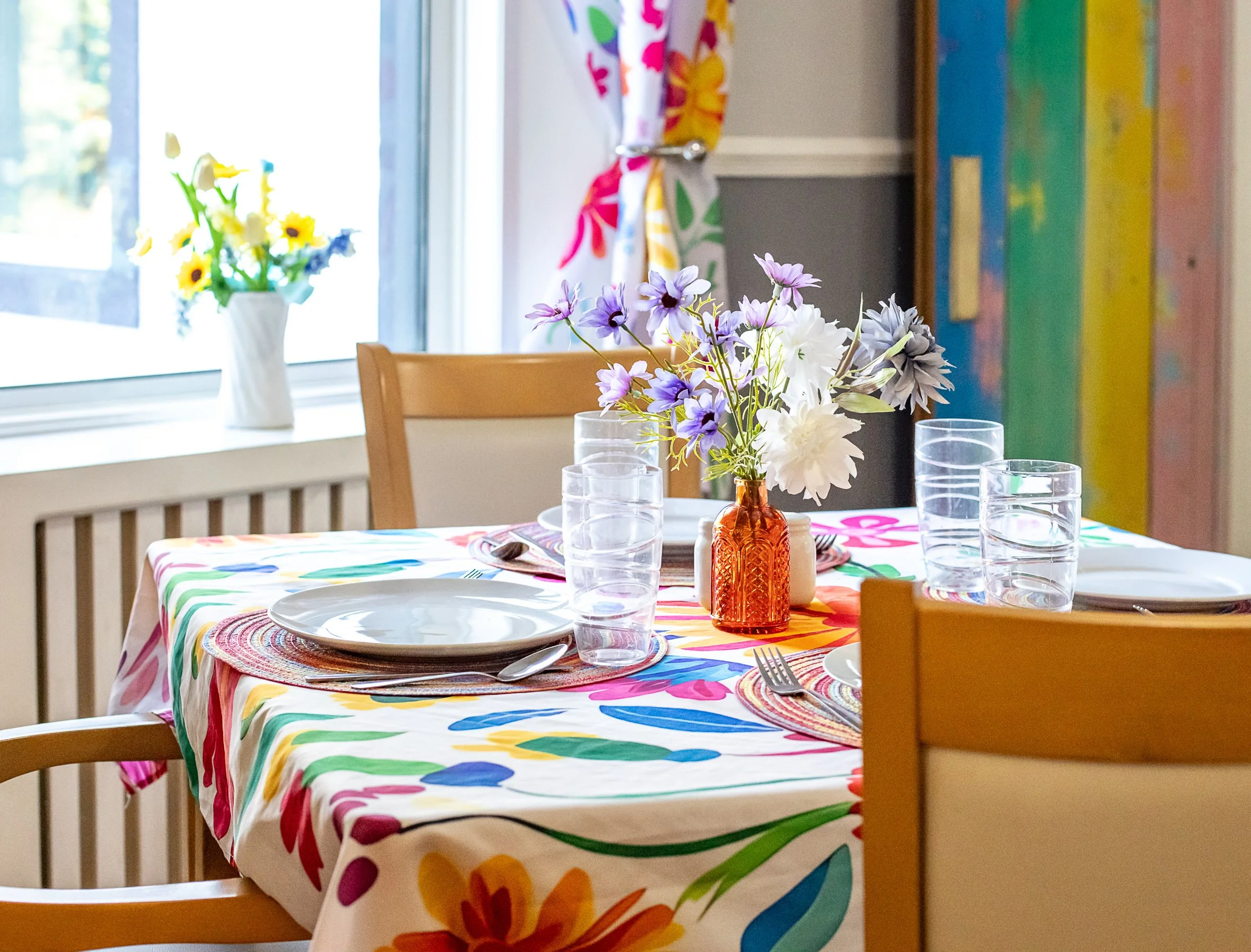 A colorful dining table set with plates, forks, and four glasses, with a floral centerpiece in an orange vase, a vase of yellow flowers near a window with a colorful curtain, and a rainbow-colored wall in the background.
