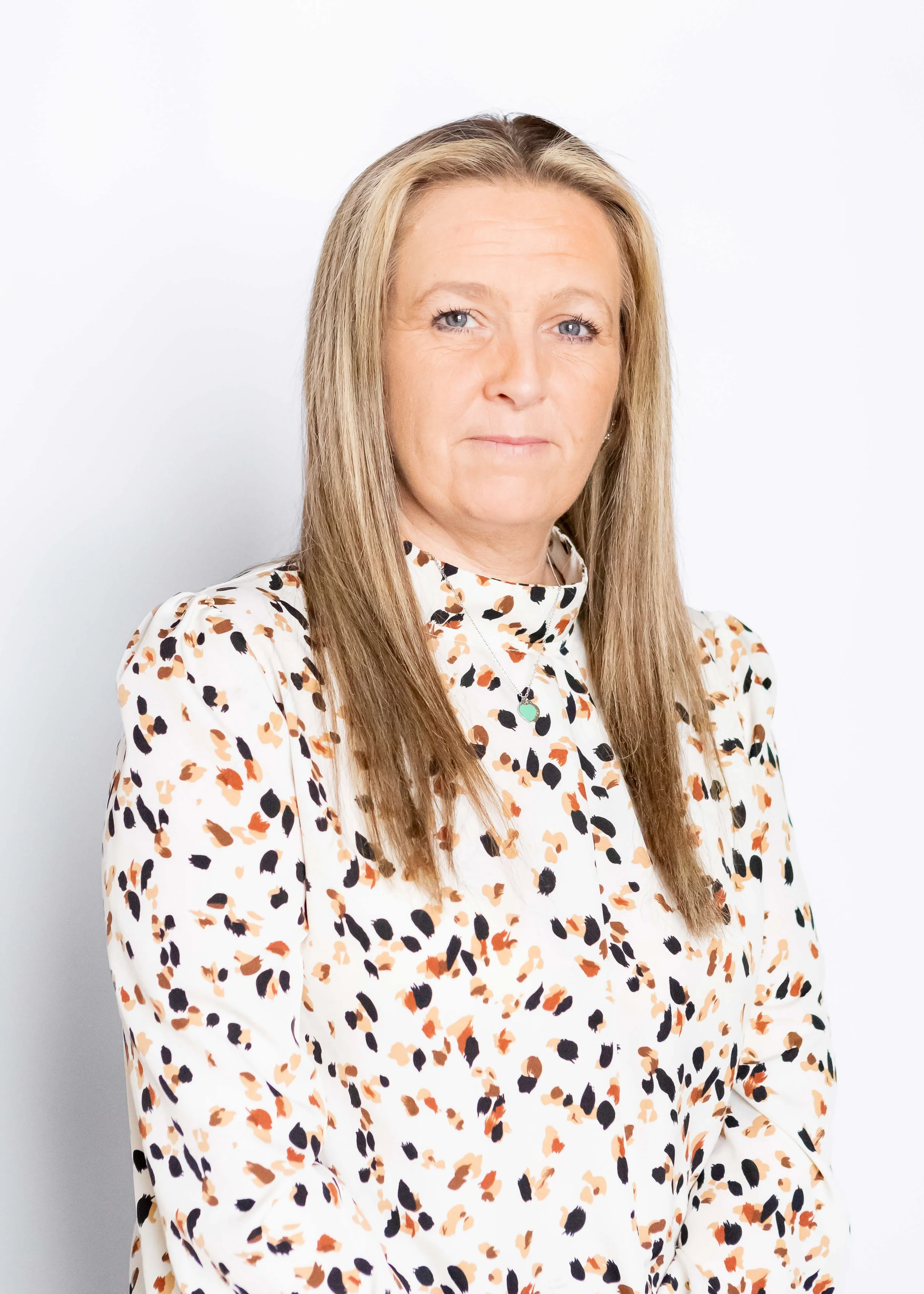 A woman with long, straight light brown hair sitting against a plain white background, wearing a white blouse with a colorful abstract pattern.