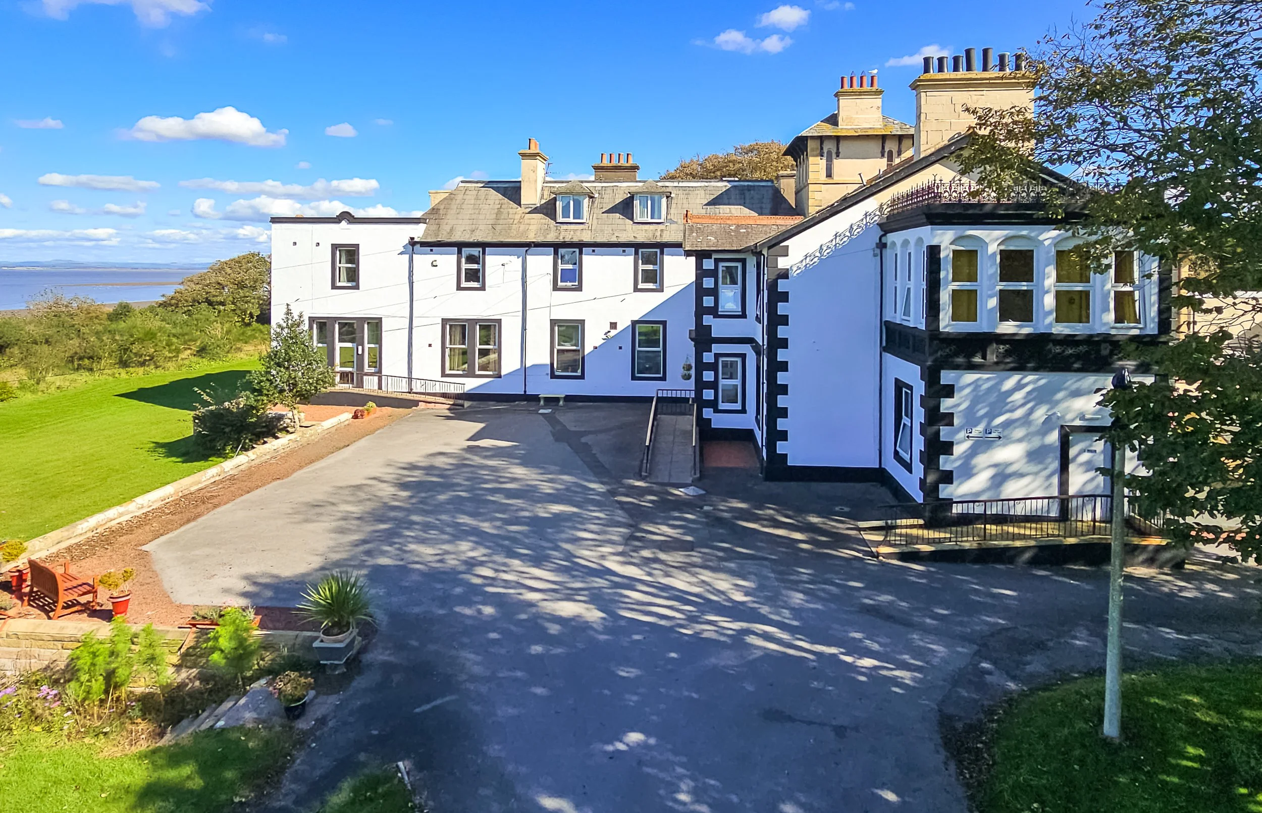 A large white multi-story house with a black and white decorative exterior, multiple windows, a driveway, and surrounding greenery under a bright blue sky.