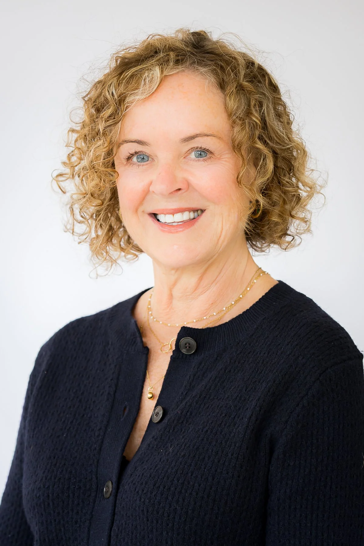 A woman with curly blonde hair and blue eyes smiling, wearing a black cardigan and gold jewelry, against a plain white background.