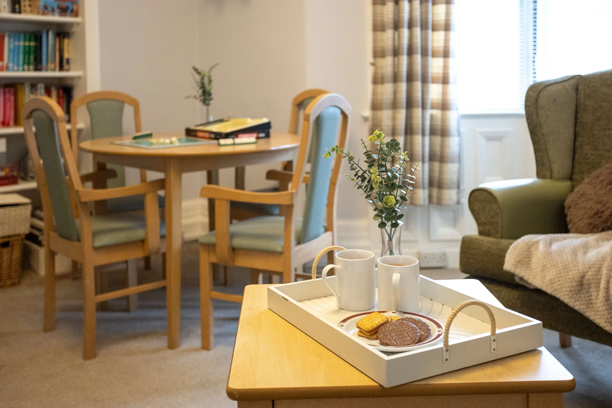A living room with a tray on a coffee table holding cups, cookies, and a flower, with a dining area and bookshelf in the background.
