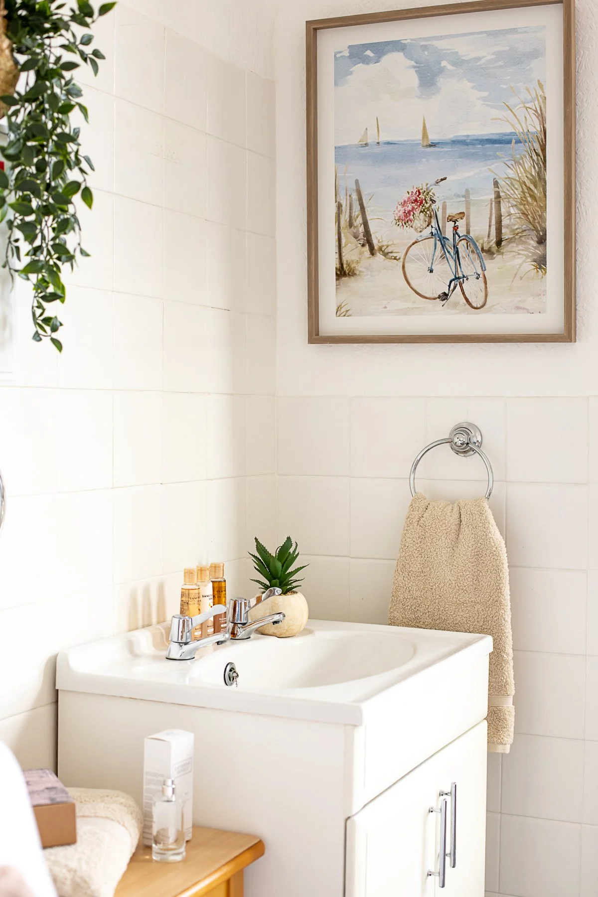 A bathroom with white tiled walls, a white vanity with a sink, and a mirror. On the vanity, there are small bottles, a small potted plant, and a decorative stone. A towel hangs on a ring next to a framed watercolor painting of a beach scene with a bi