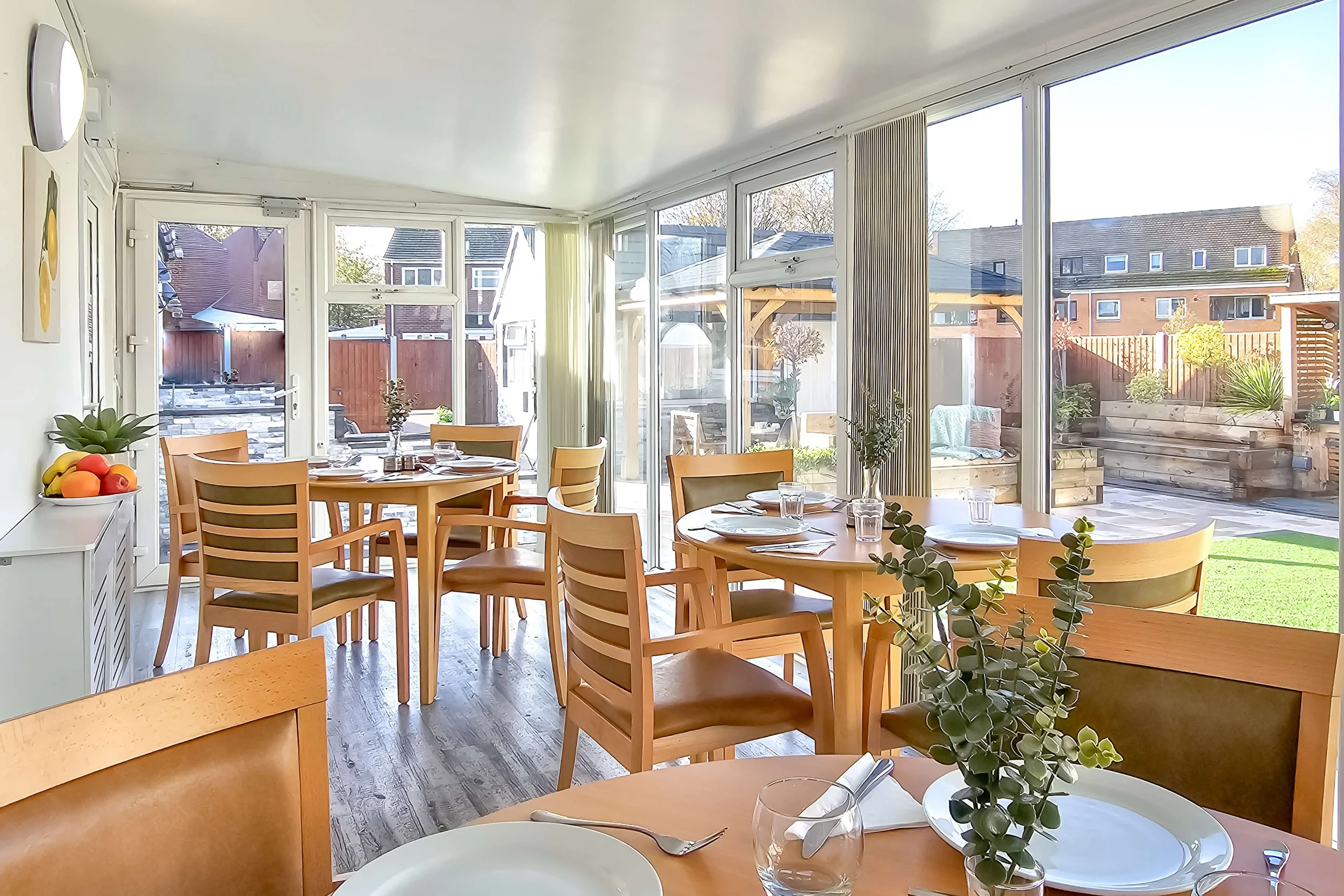 Sunlit dining room with wooden tables and chairs, set with plates, glasses, and cutlery, adjacent to a backyard with a patio and garden.