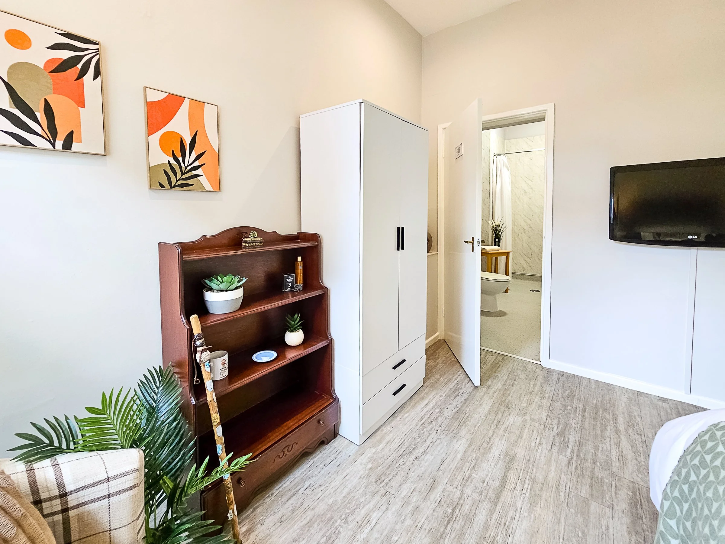 A bedroom with a white wall, a wooden bookshelf with plants and decorative items, a white wardrobe, a small doorway to a bathroom, a mounted flat-screen TV, part of a bed with a patterned blanket, and framed abstract artwork on the wall.