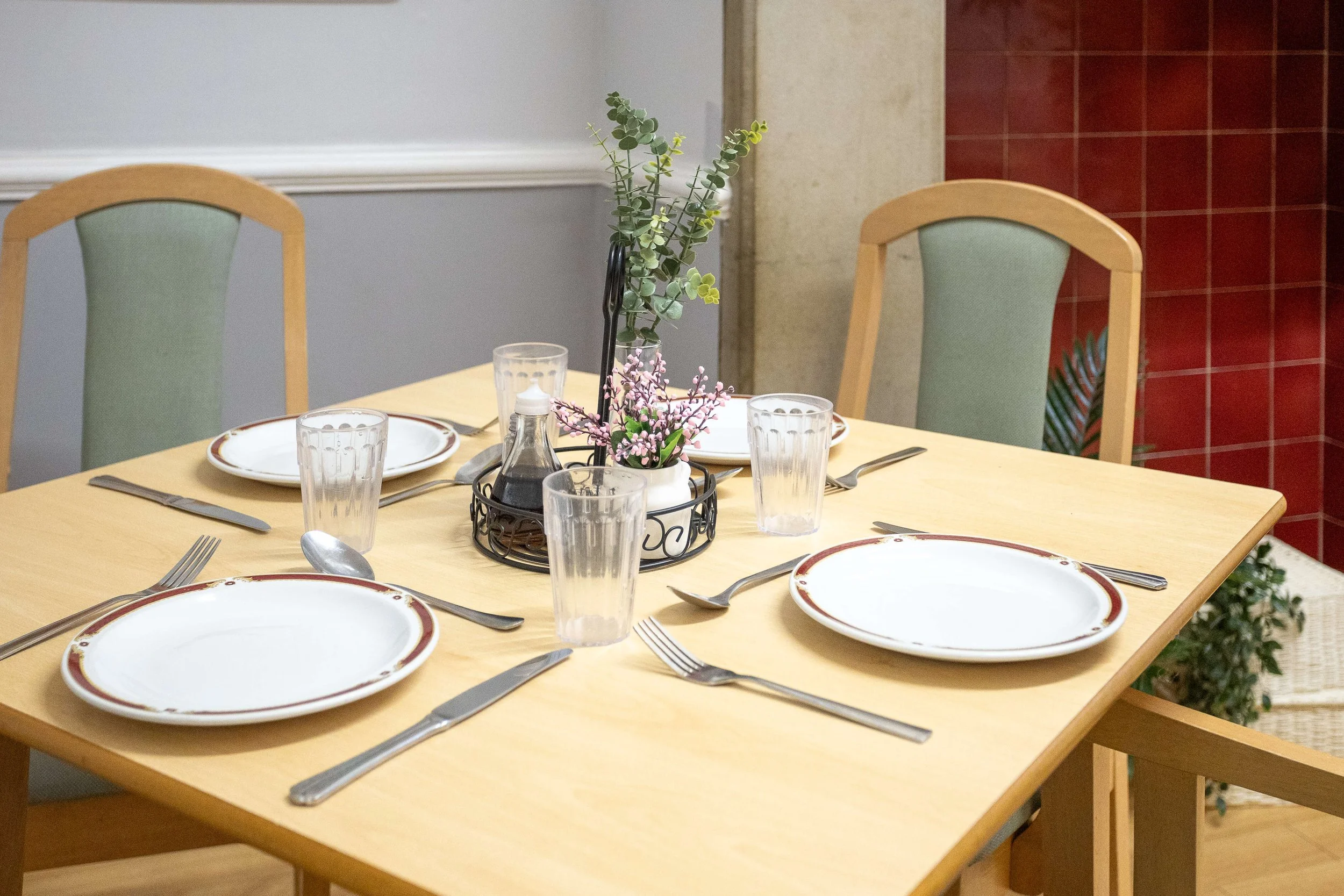 A neatly set dining table with four plates, four glasses, and cutlery, featuring a centerpiece with greenery and pink flowers, in a cozy restaurant setting.