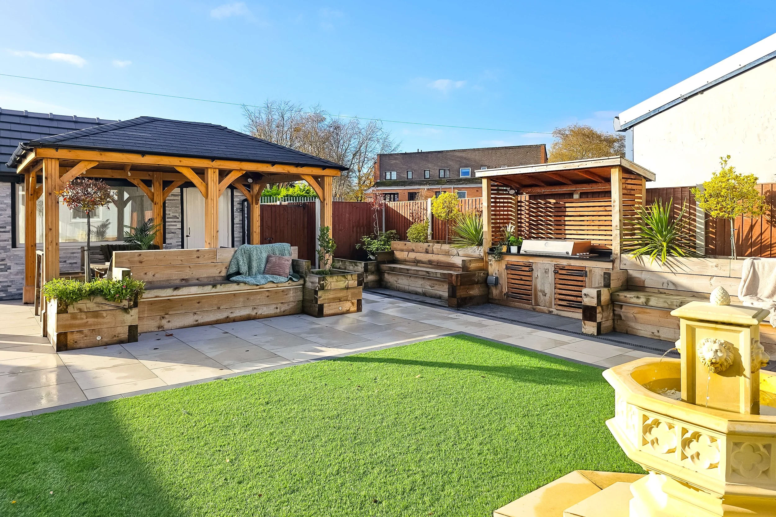 Backyard patio with wooden pergola, outdoor seating, potted plants, a small fountain, and a lawn under clear blue sky.