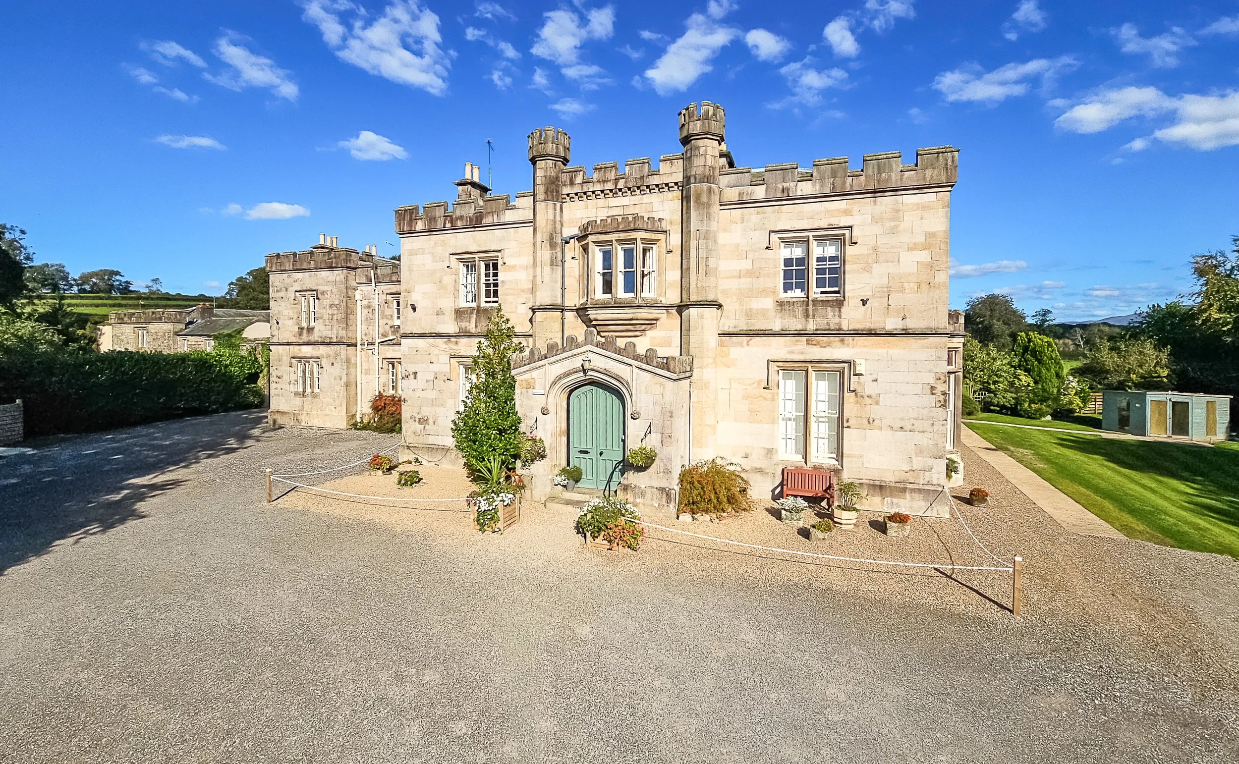 A historic castle with a stone facade, multiple small turrets, and a green arched door, surrounded by a gravel courtyard, potted plants, and a grassy garden with trees under a blue sky with scattered clouds.