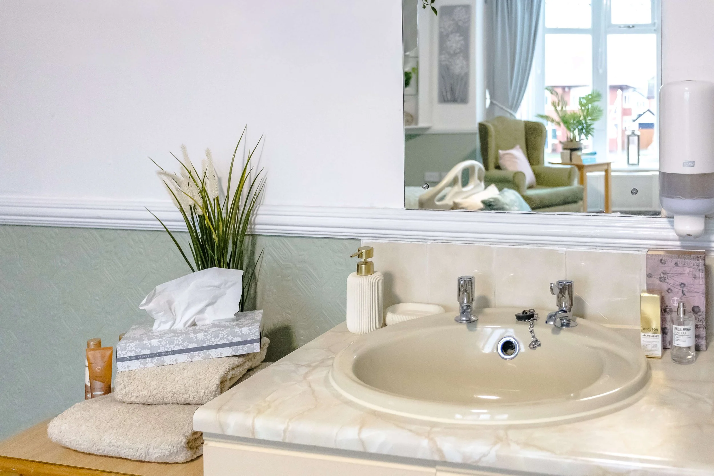 Bathroom sink with beige countertop, soap dispenser, tissue box, plant, mirror, and various toiletries. Reflection in the mirror shows an armchair, side table, window with curtains, and a view outside.