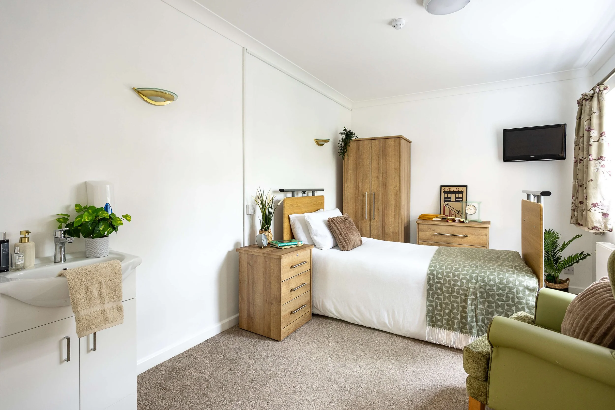 A clean, well-lit hotel room with a single bed, a bedside table, a wardrobe, a small dresser with decorative items, a wall-mounted TV, a green armchair, and an adjacent sink area with a plant and toiletries.