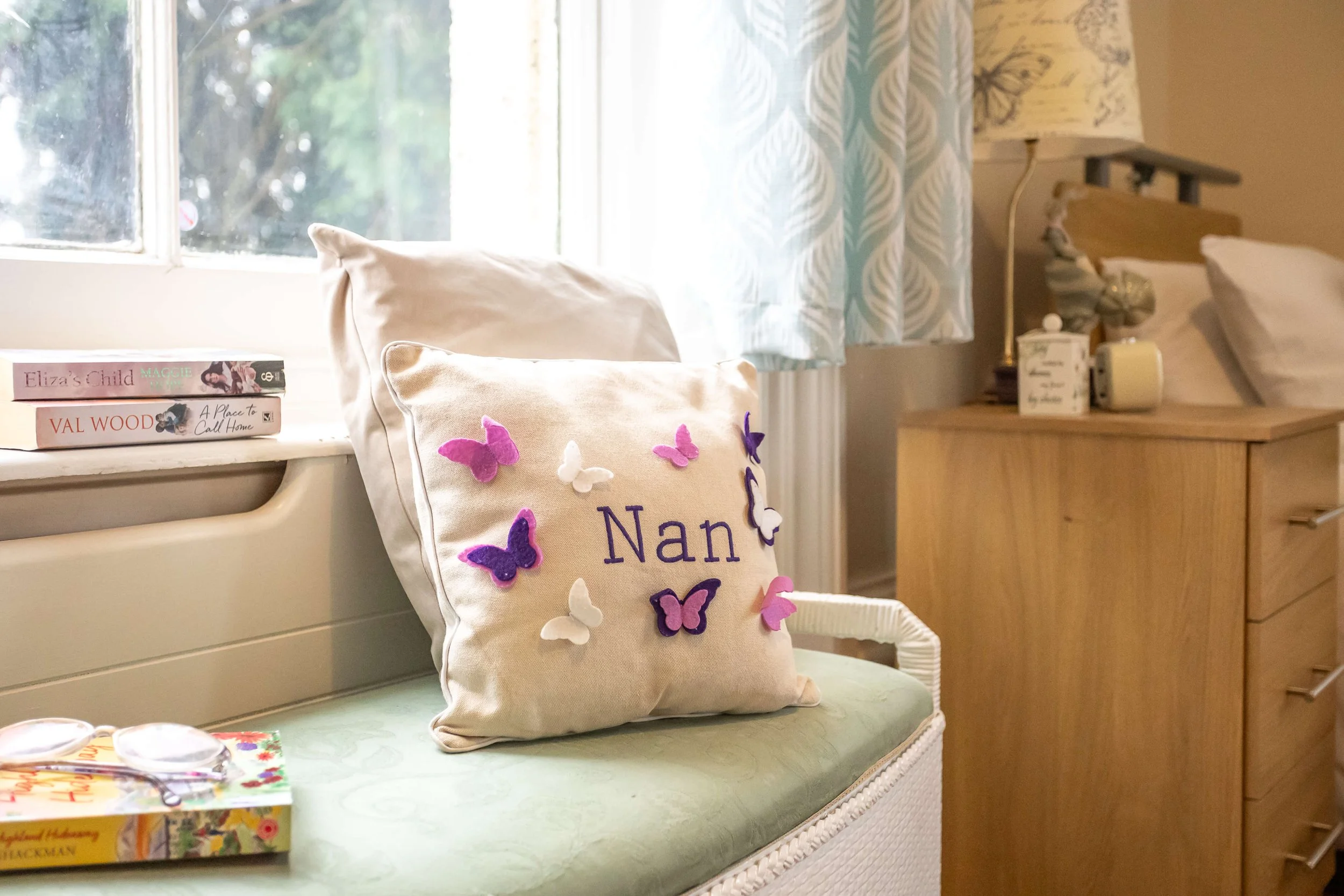 Decorative pillow with butterflies and the name 'Nan' on it, placed on a light green upholstered window seat in a cozy bedroom with a wooden dresser, books, and a window with light blue patterned curtains.