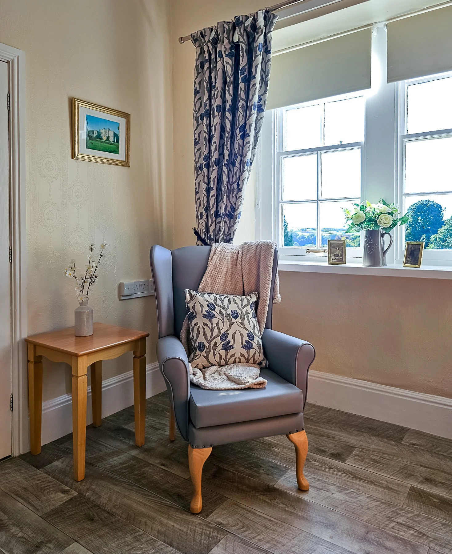 A cozy reading nook with a blue wingback chair, cushion, and blanket near a window, with a small wooden side table, a vase with flowers, and framed photos on the windowsill.