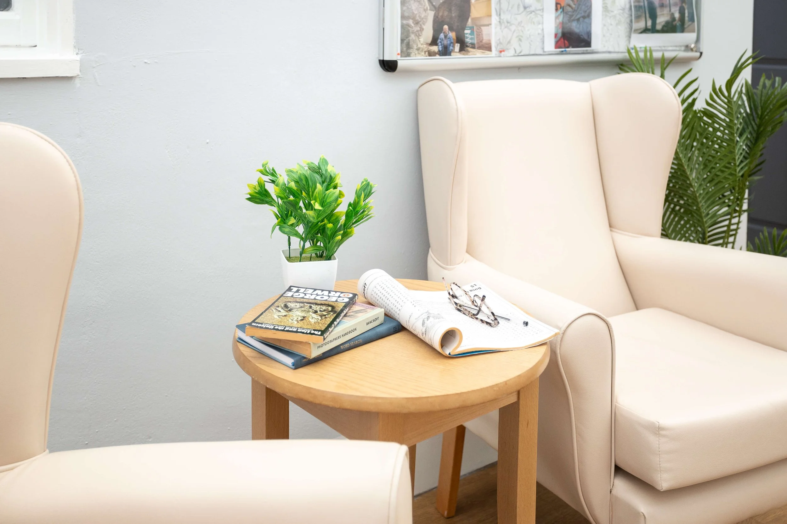 A cozy room corner with a round wooden table holding books, a rolled-up magazine, eyeglasses, and a potted green plant. Two off-white armchairs are positioned around the table, with a bulletin board and a tall plant with large leaves in the backgroun