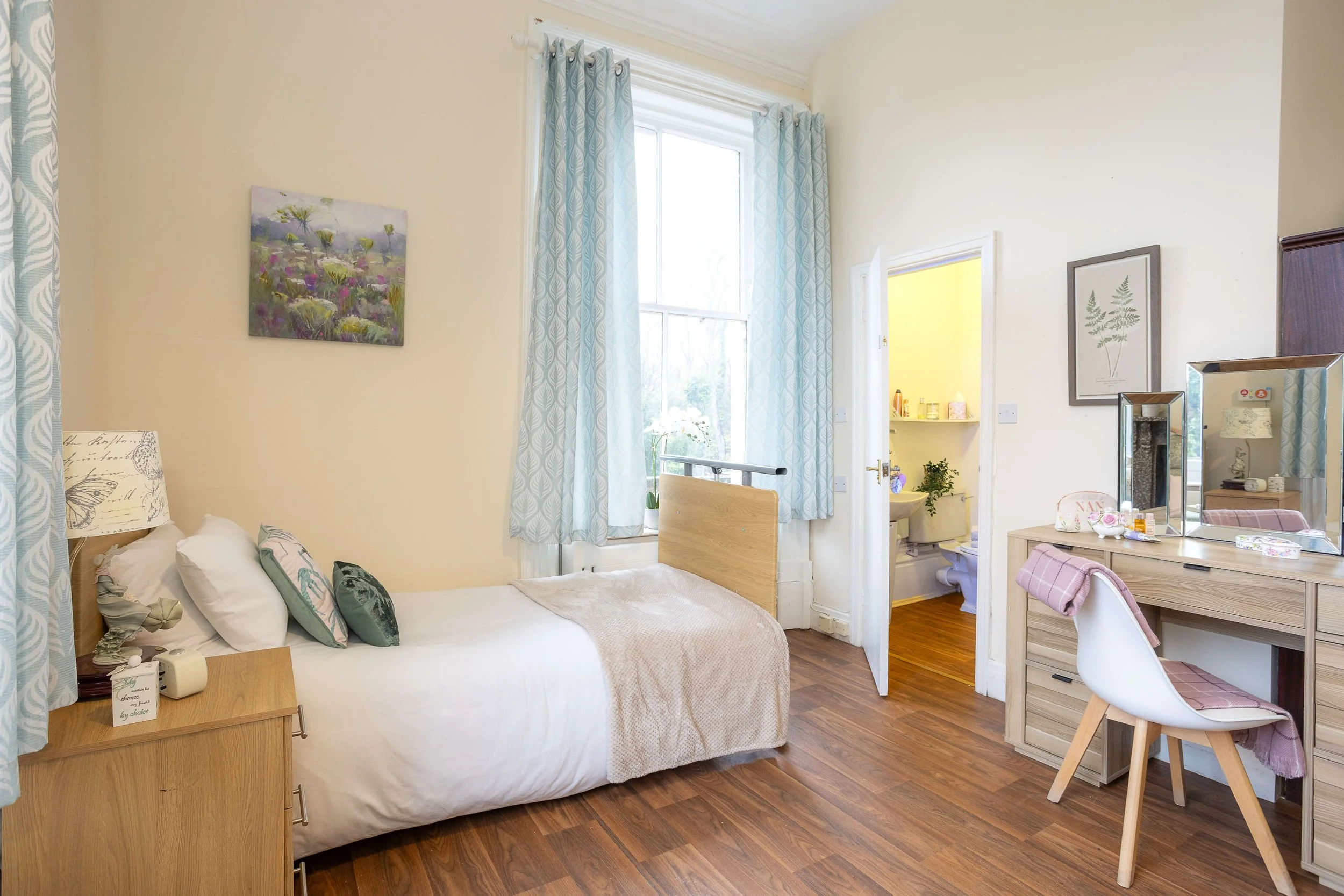 A cozy bedroom with a single bed, light-colored wooden furniture, a window with light blue curtains, a small vanity with a mirror, and an en-suite bathroom visible through an open door.