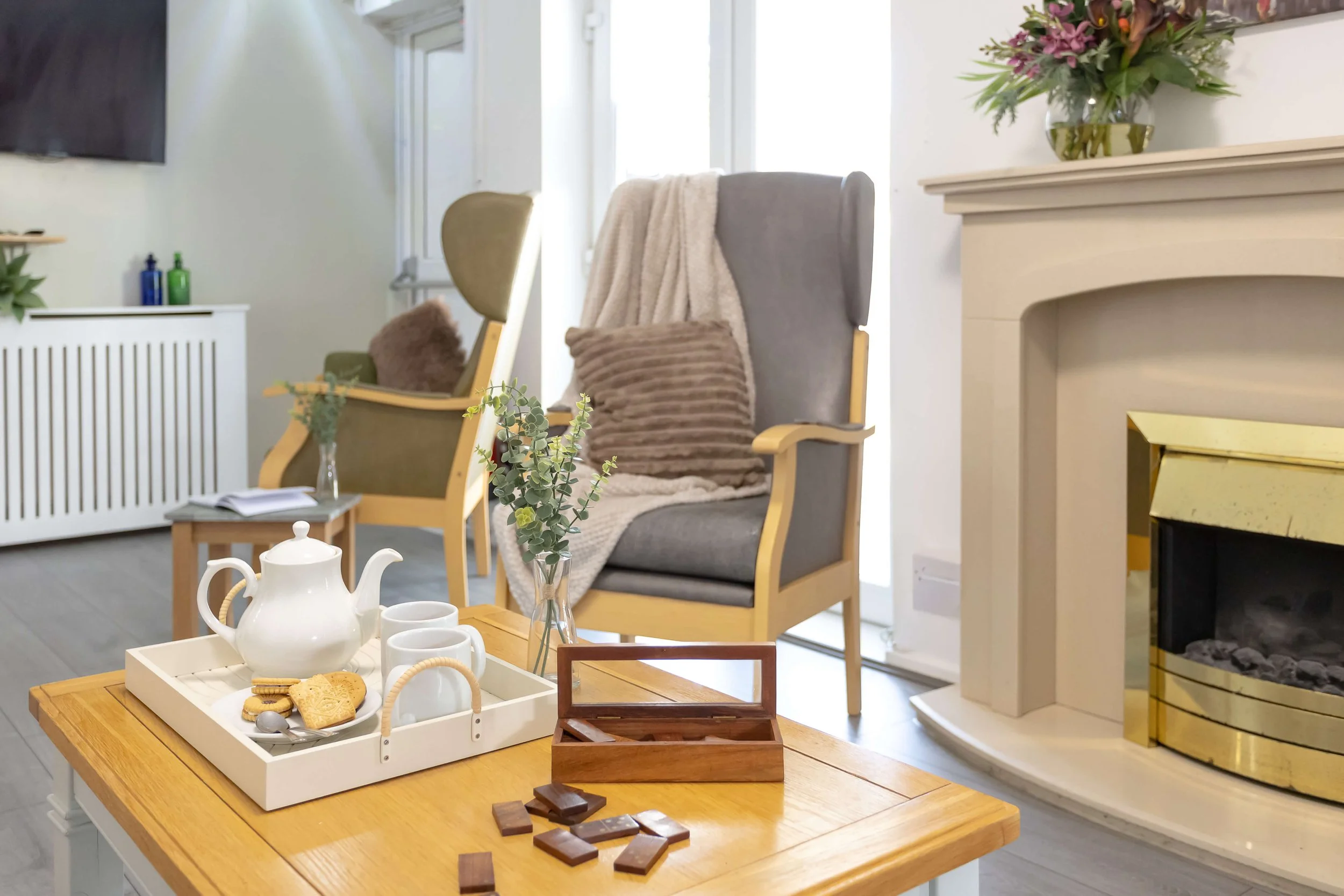 Cozy living room with two armchairs, one with a brown pillow and blanket, a wooden coffee table with a teapot, cups, and cookies, a side table with books and a vase of eucalyptus, a fireplace with a golden trim, and a vase of pink flowers on the mant