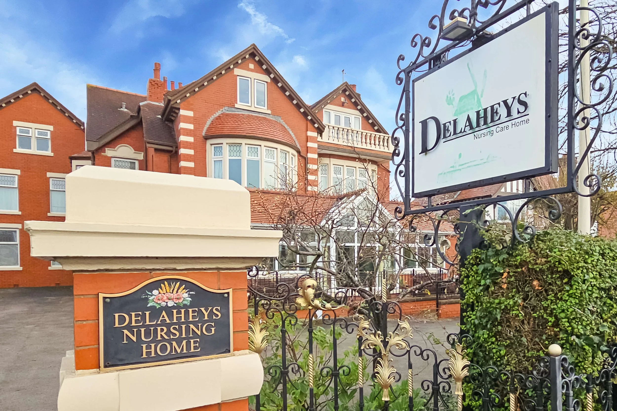 Entrance of Delahey's Nursing Home with signboards, a red brick building with white window frames, and a decorative wrought iron fence.
