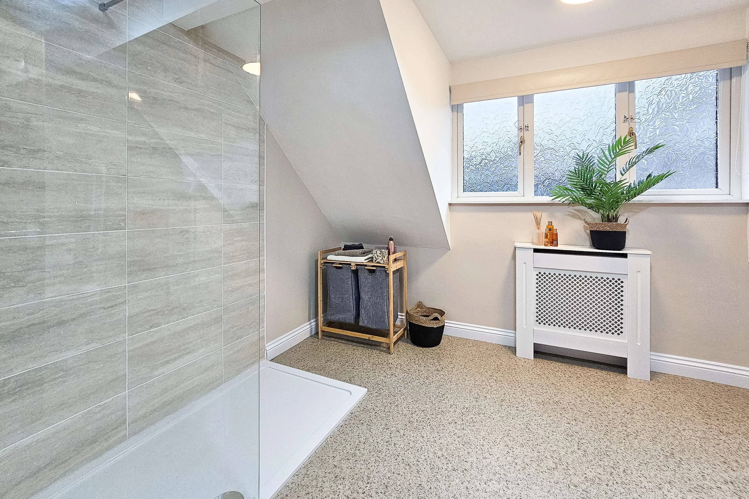 Bathroom corner with a glass shower, beige tiled wall, white radiator cover, potted plant, window, and small storage stand with toiletries.
