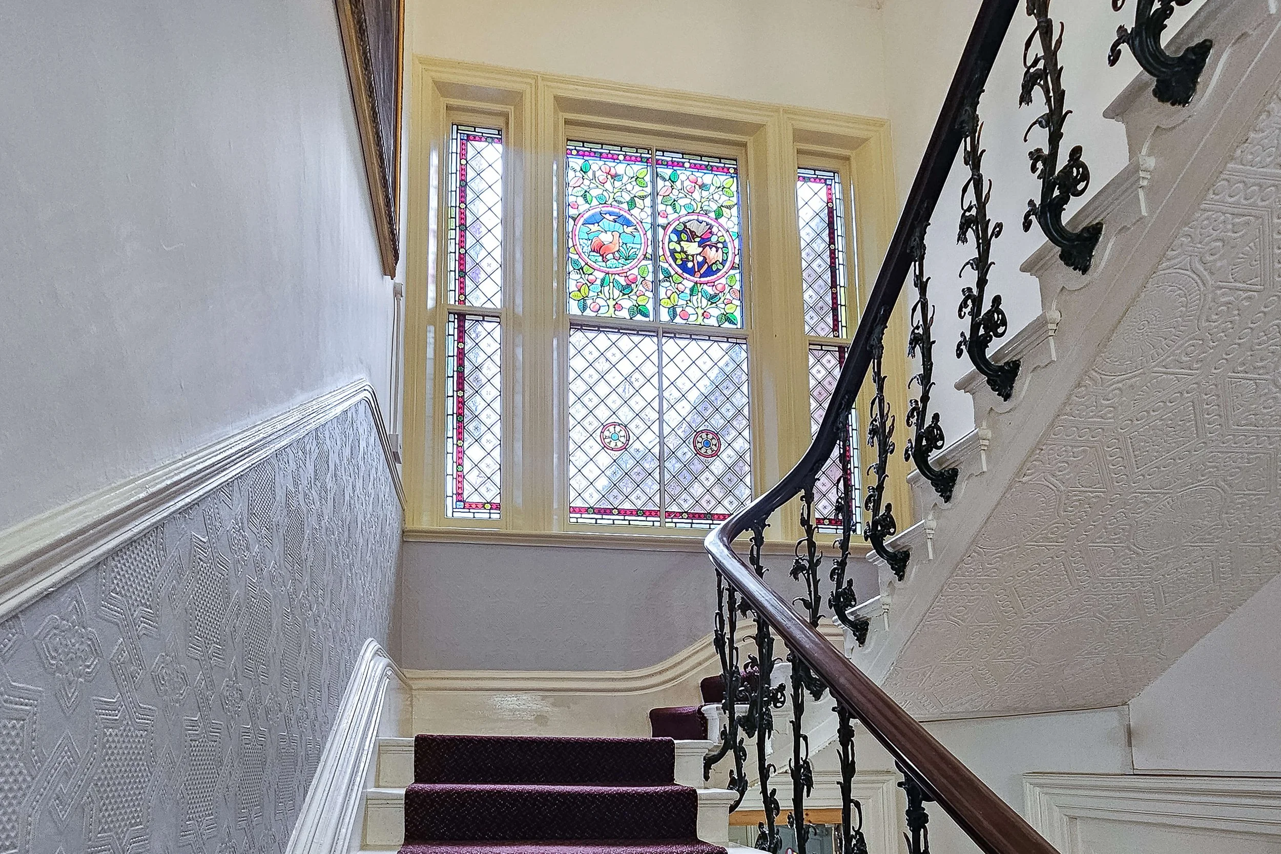 Interior view of a staircase with a decorative handrail and a large stained glass window at the top, featuring various colored sections and patterns.