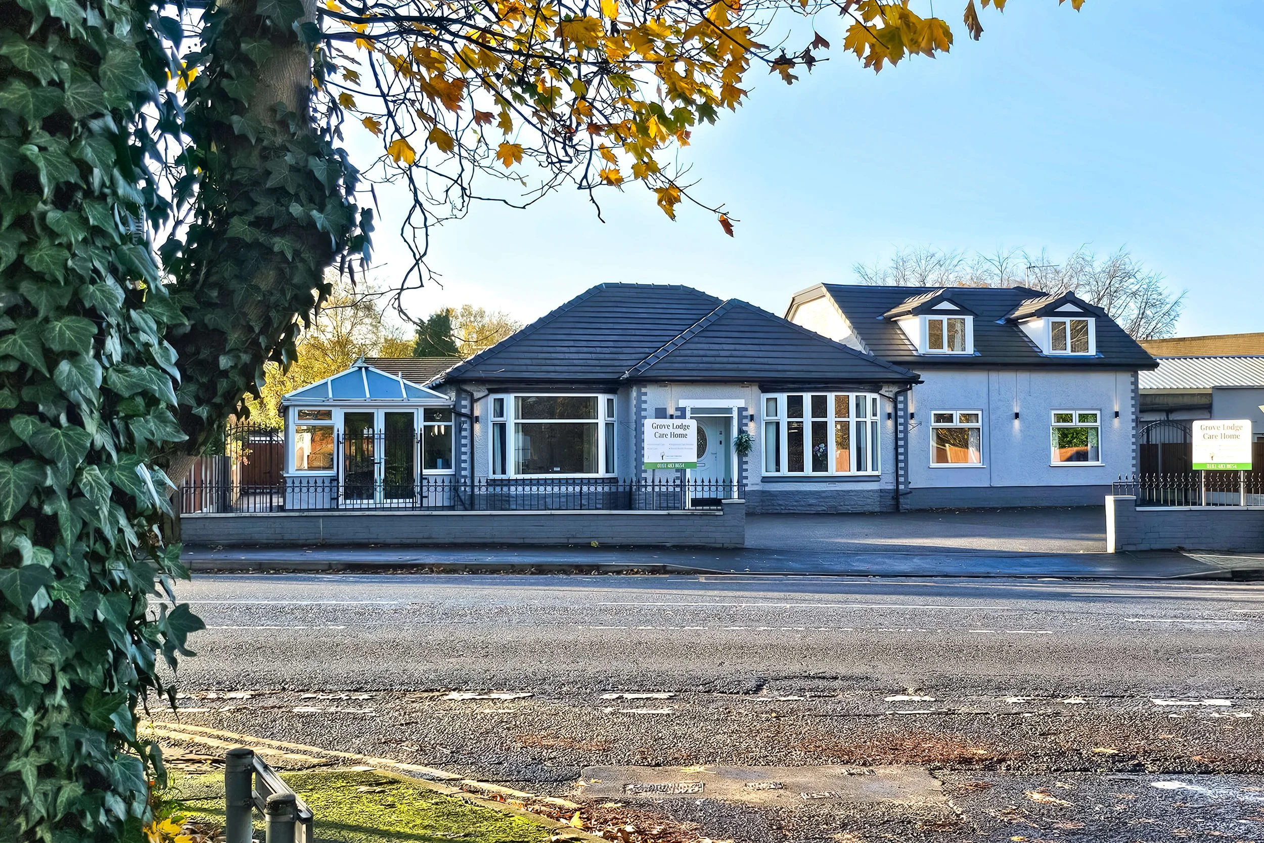 A residential building with a dark tiled roof, large windows, and a small conservatory, situated behind a fence along a street.