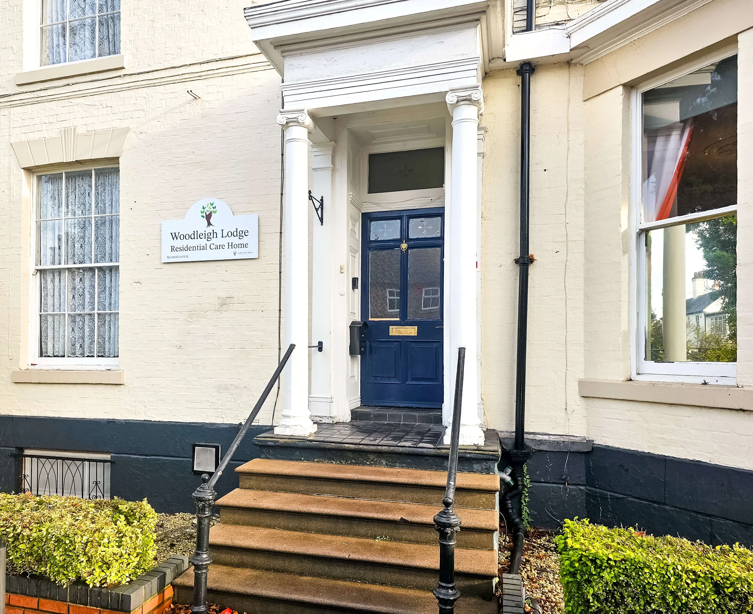 Exterior view of a residential care home named Woodleigh Lodge, with a blue front door, white pillars, a ramp, and stairs, with windows on either side and greenery in front.