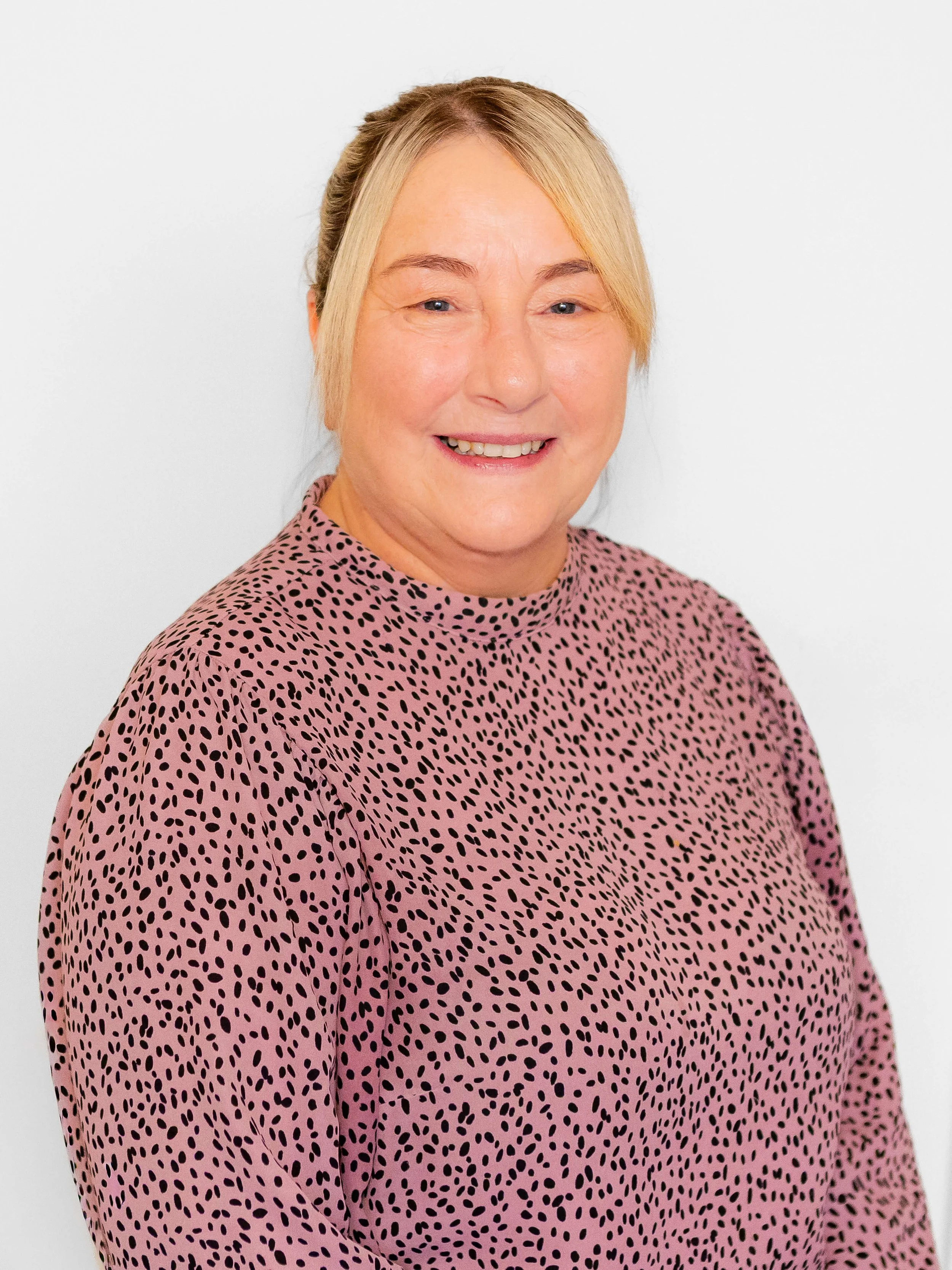A middle-aged woman with blond hair pulled back, smiling, wearing a pink and black spotted blouse, standing against a plain white background.