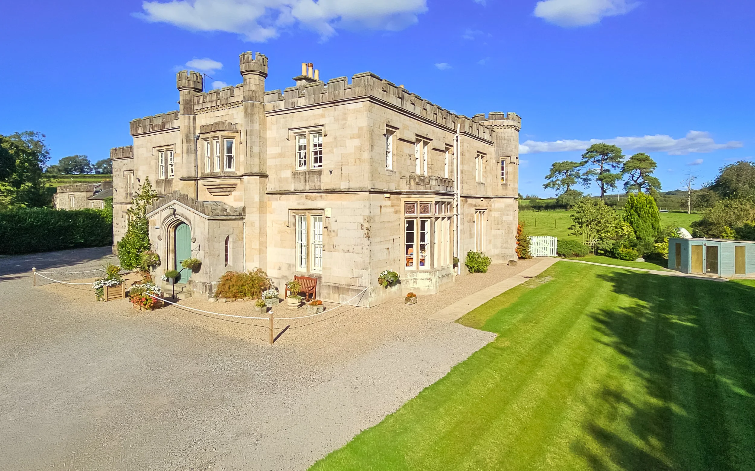 A large stone castle with battlements and multiple windows in a scenic rural landscape with green grass, trees, and a partly cloudy blue sky.