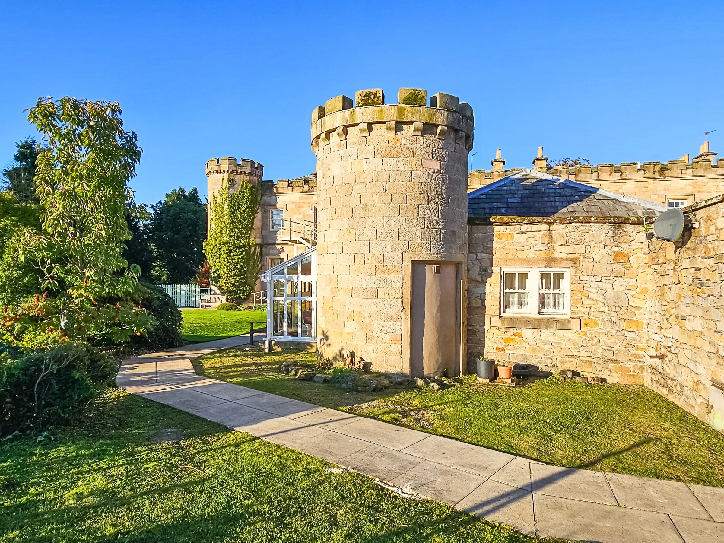 A stone castle-style building with round towers, small windows, and a satellite dish on the wall, surrounded by a well-maintained garden and a curved sidewalk, under a clear blue sky.