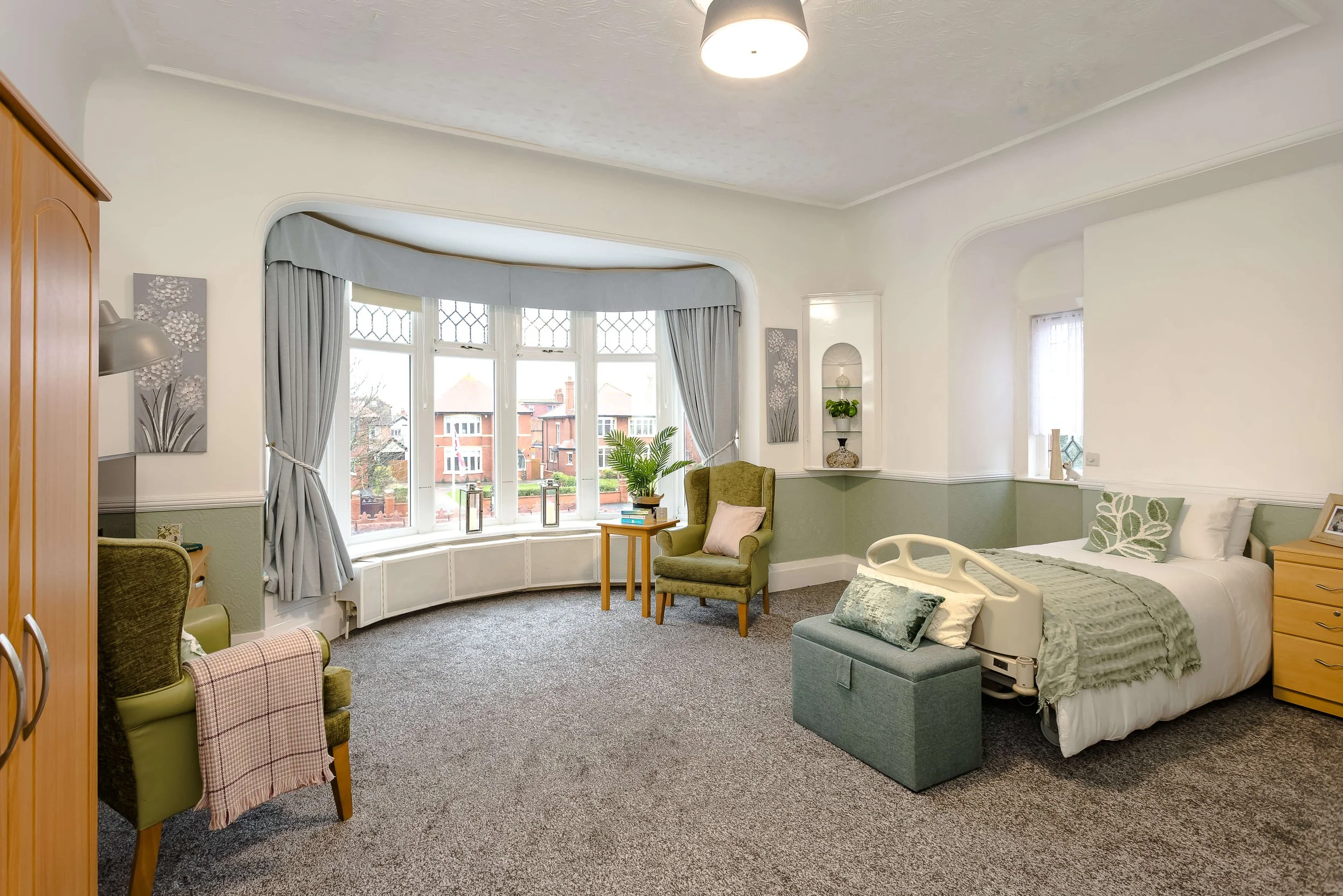 A brightly lit bedroom with a large bay window, green armchair, bed with pillows, and various decorative elements