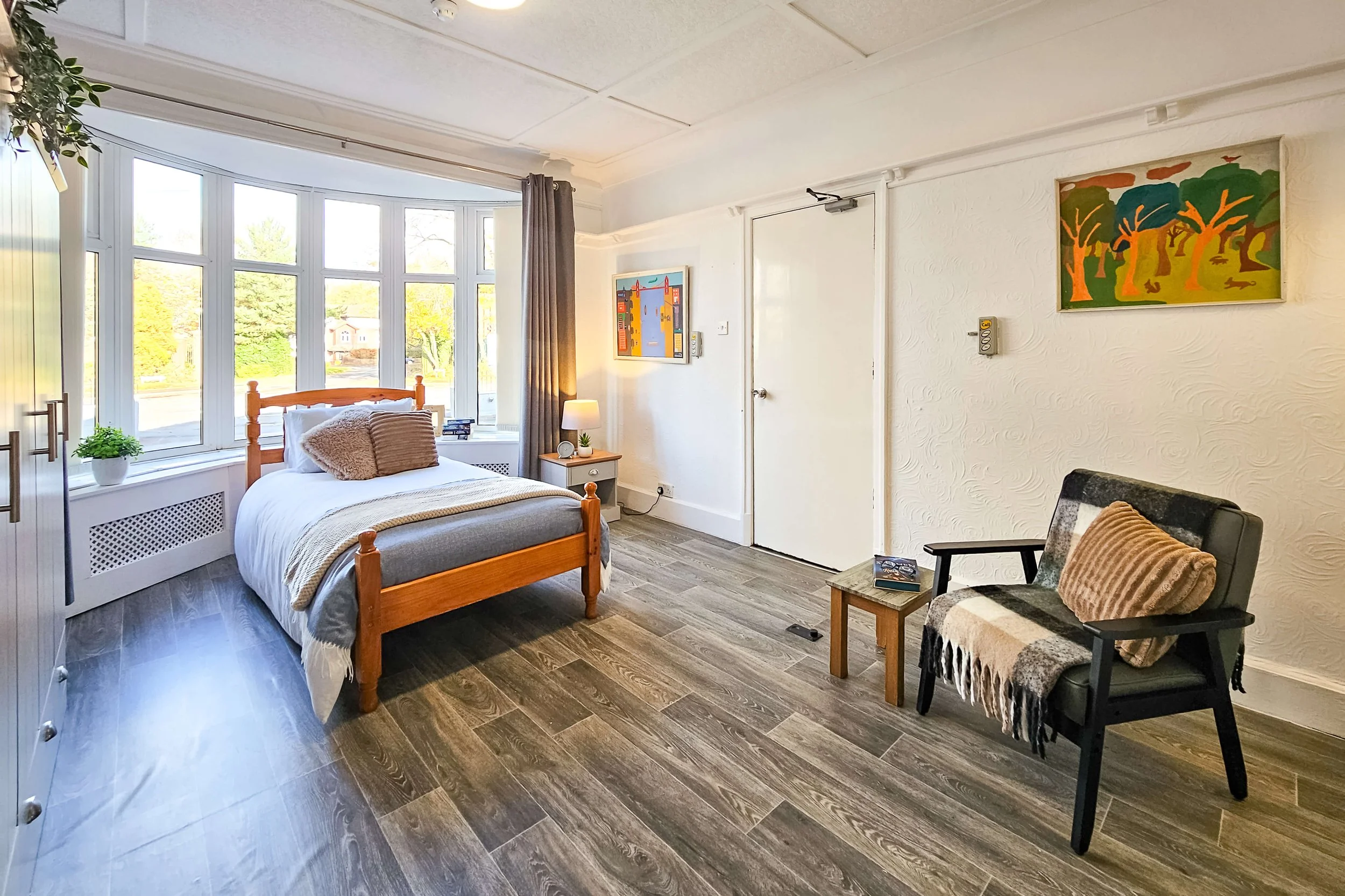 A cozy bedroom with a large window, a bed with white bedding and brown pillows, a wooden nightstand with a lamp, framed artwork on the walls, a black chair with a plaid throw and pillow, and wood flooring.