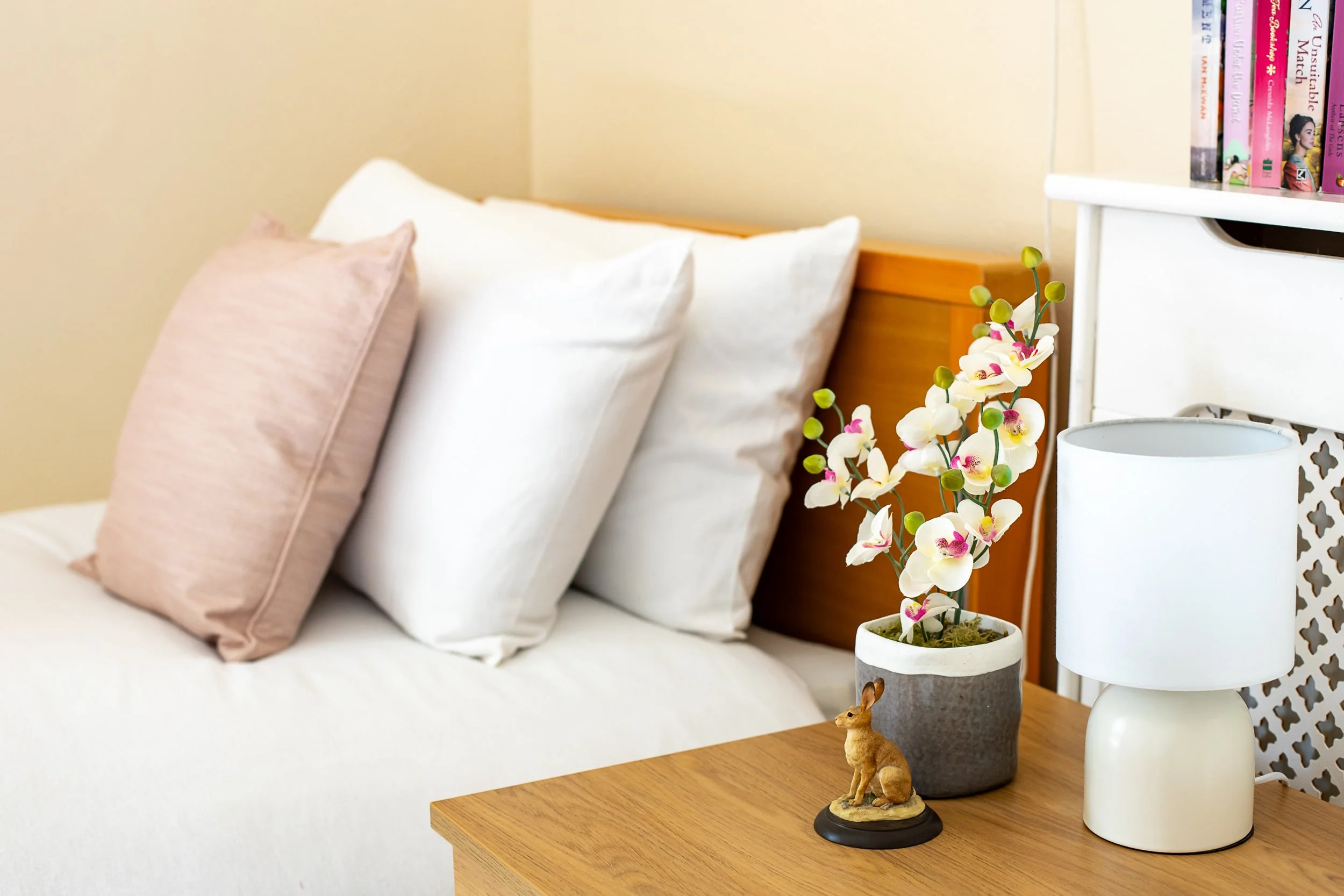 A bedroom with a bed, white bedding, pink and white pillows, a wooden nightstand with a potted orchid, a lamp, and a bookshelf.